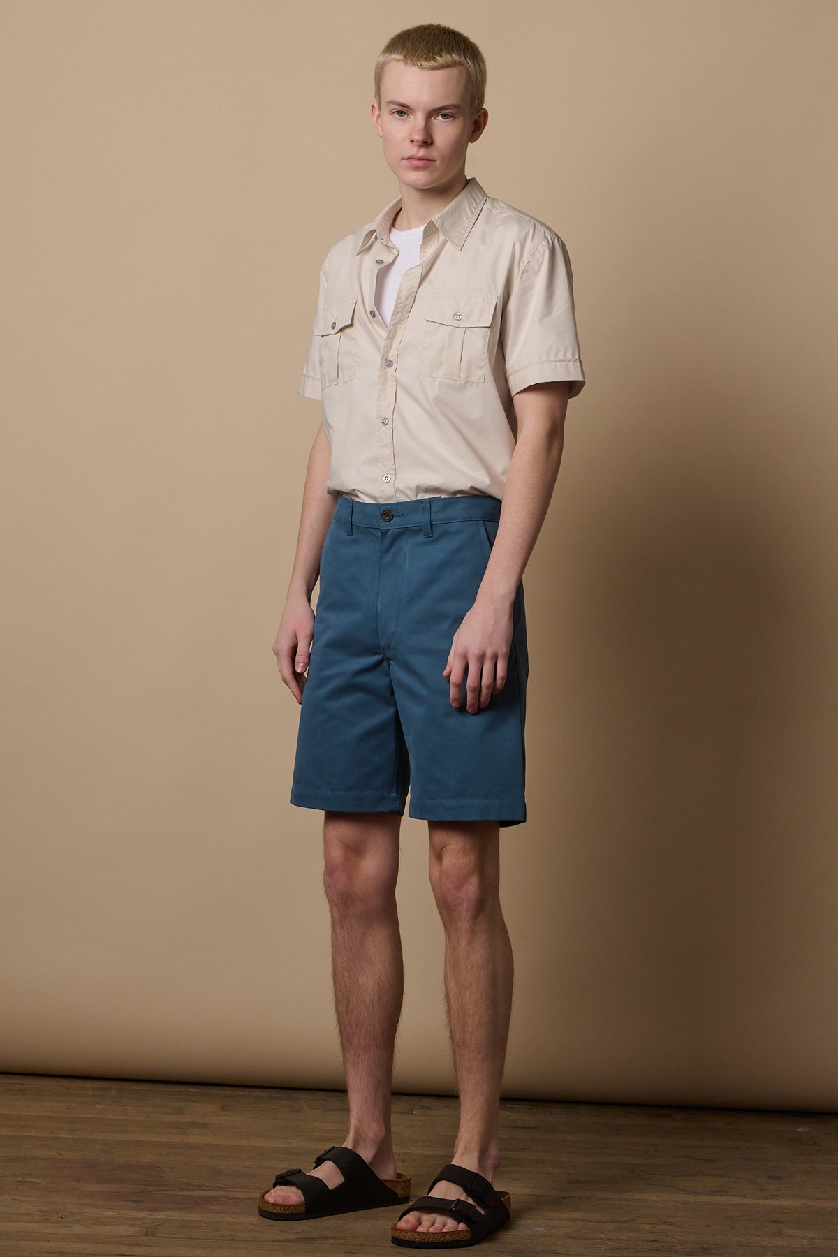 Full body image of white male with short blonde hair wearing shirt tucked into classic shorts in RAF blue and Birkenstock sandles