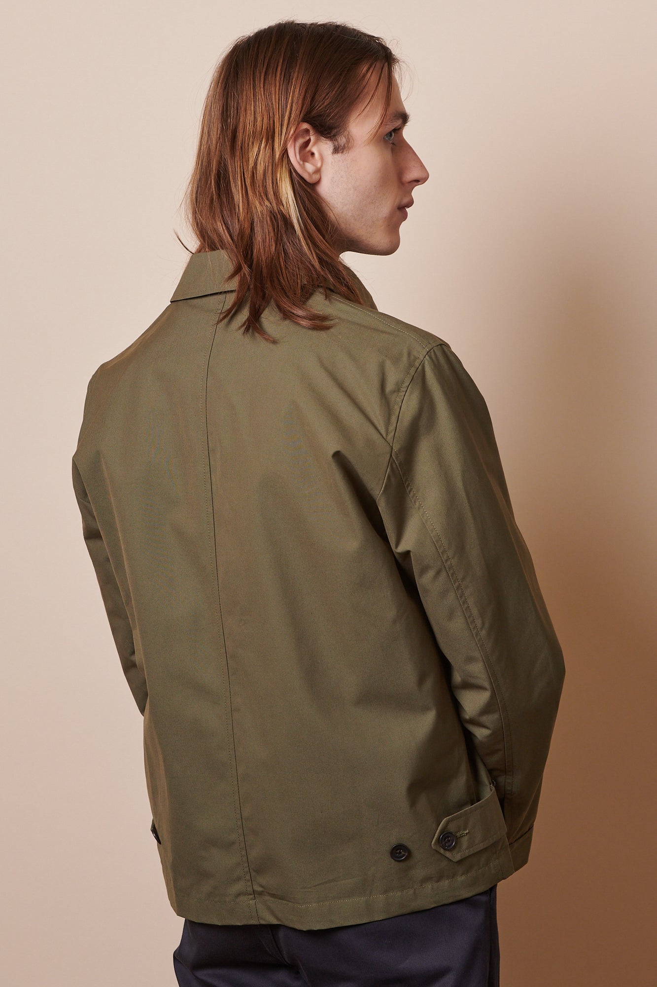 The back of male with shoulder length hair wearing collared Harrington in olive, buttons for adjusting the hem width