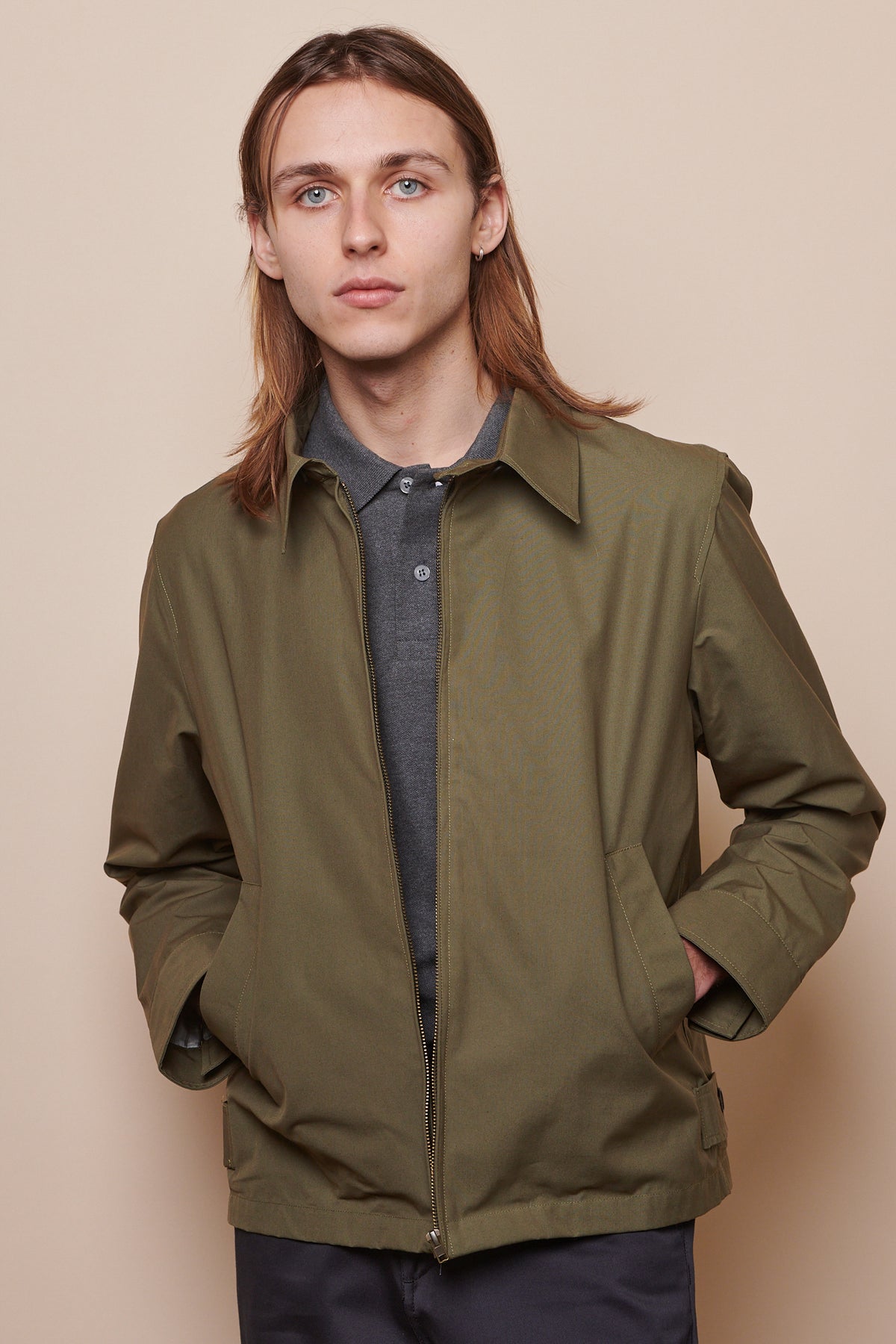 Thigh up image of male wearing unzipped collared Harrington jacket in olive with both hands in front pockets