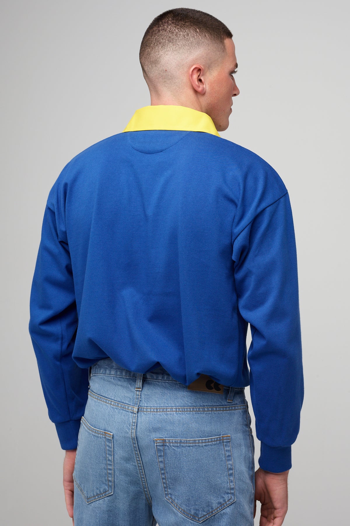 
            White, brunette male facing away, wearing blue rugby shirt, contrast yellow collar, tucked into blue jeans,