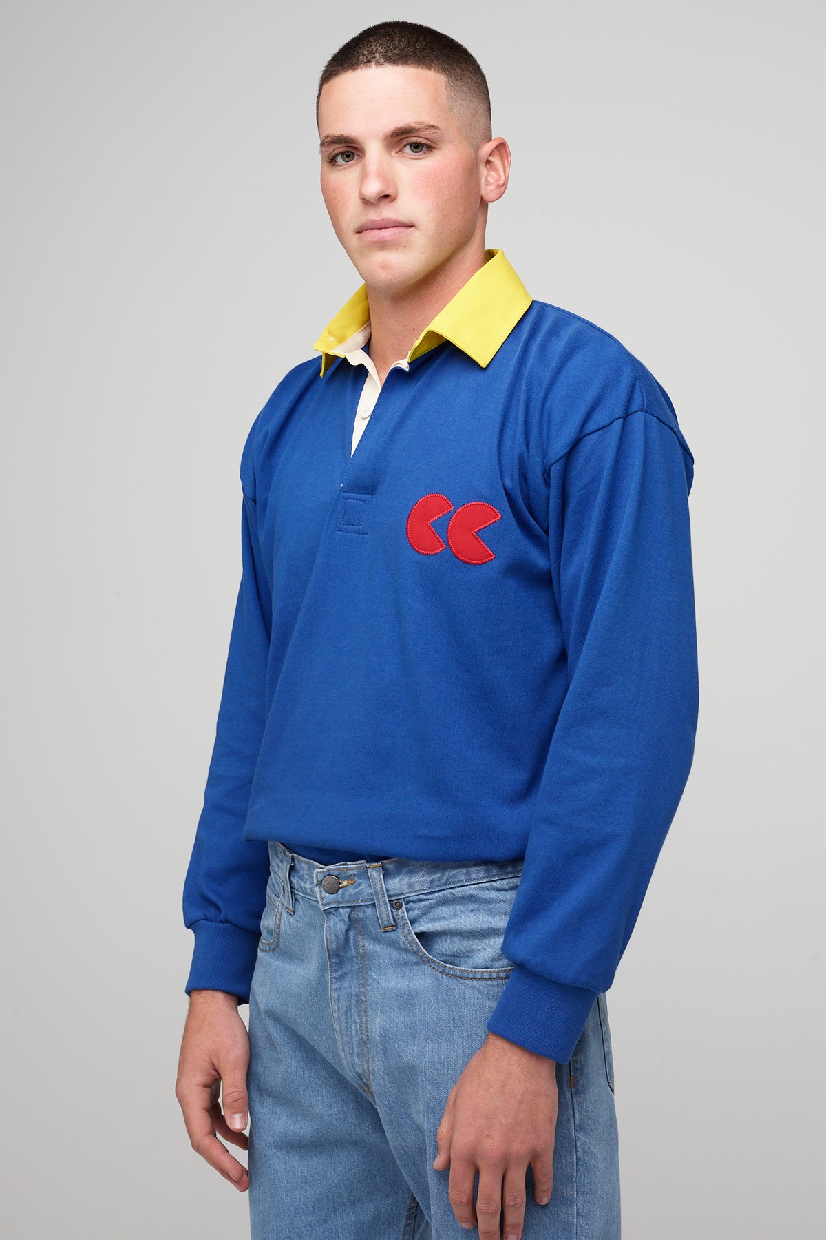 
            White, brunette male wearing blue long sleeve rugby shirt, contrast yellow collar, red CC logo badge tucked into blue jeans