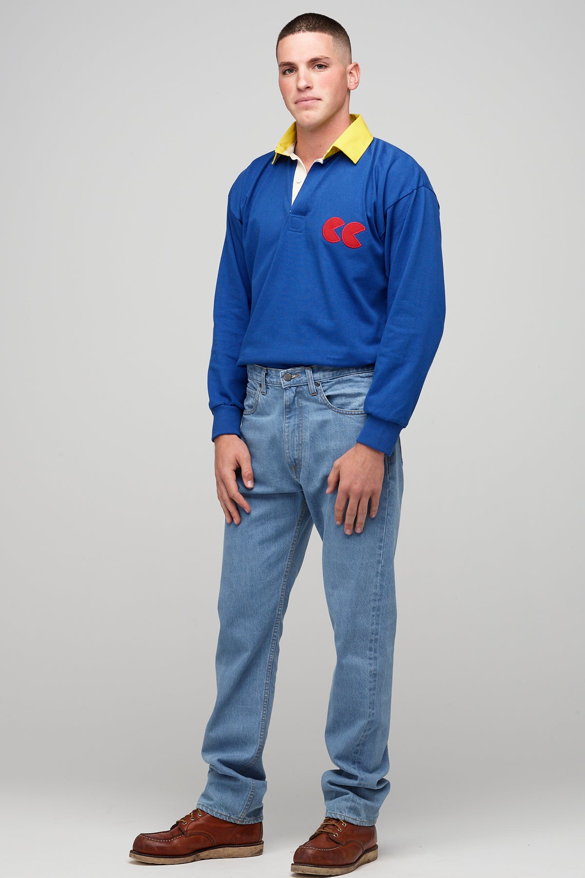 
            White, brunette male wearing long sleeve blue rugby shirt, contrast yellow collar, red CC logo badge, styled, tucked tucked into denim jeans with brown shoes