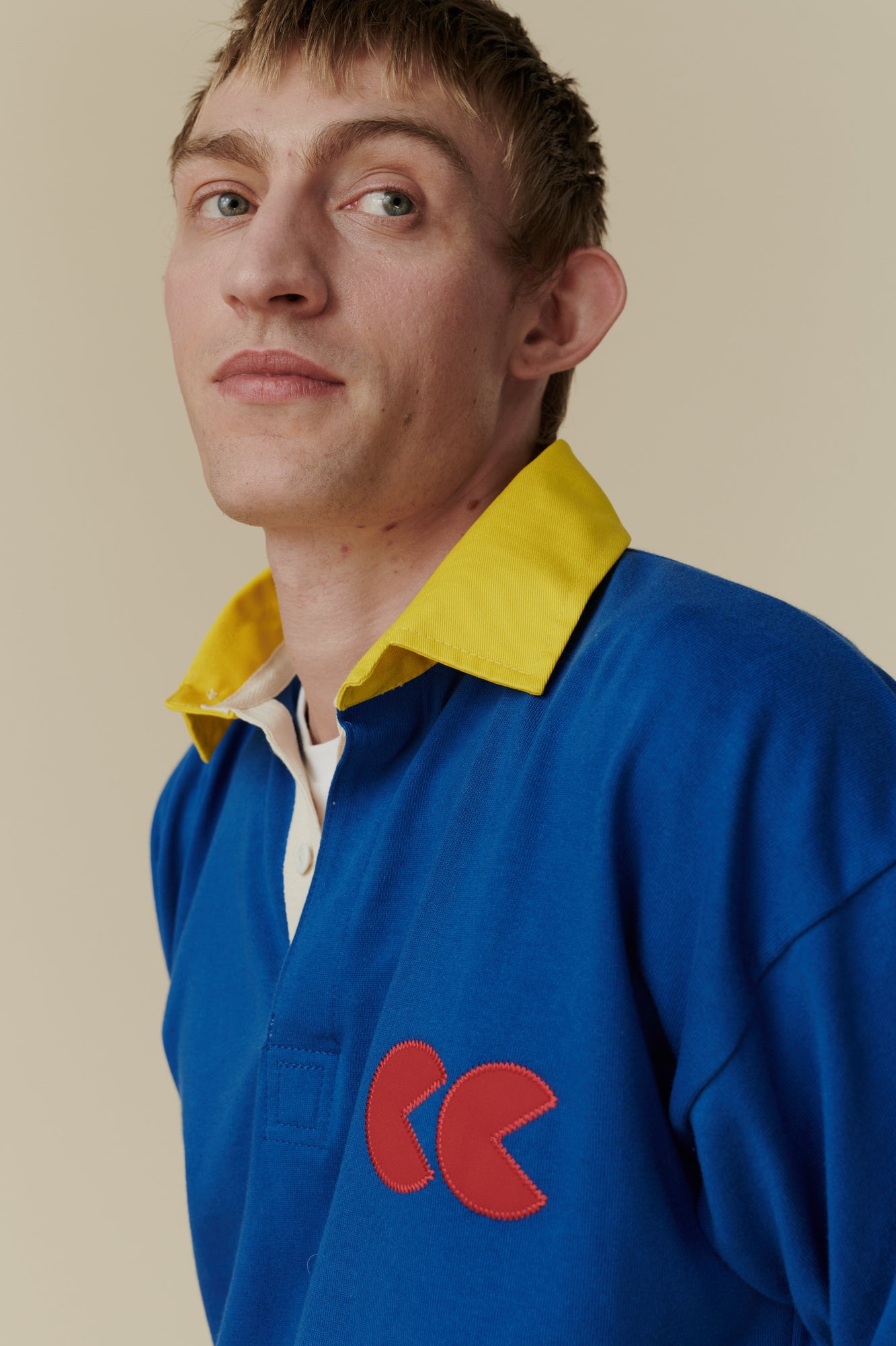 Close up portrait of white male wearing long sleeve blue rugby shirt with contrast yellow collar and red CC logo badge on chest, worn over plain white t shirt.