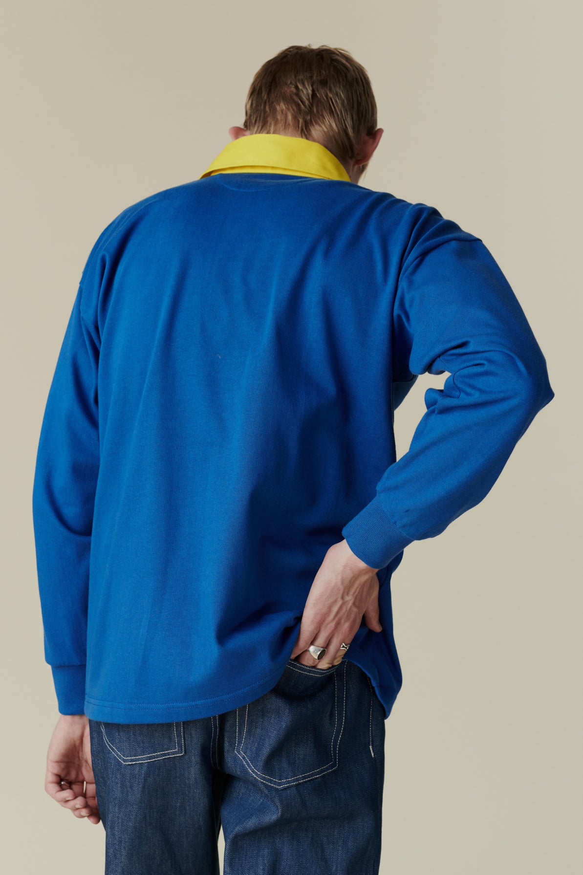The back of white male wearing long sleeve blue rugby shirt with contrast yellow collar and red CC logo badge on chest, worn over plain white t shirt.