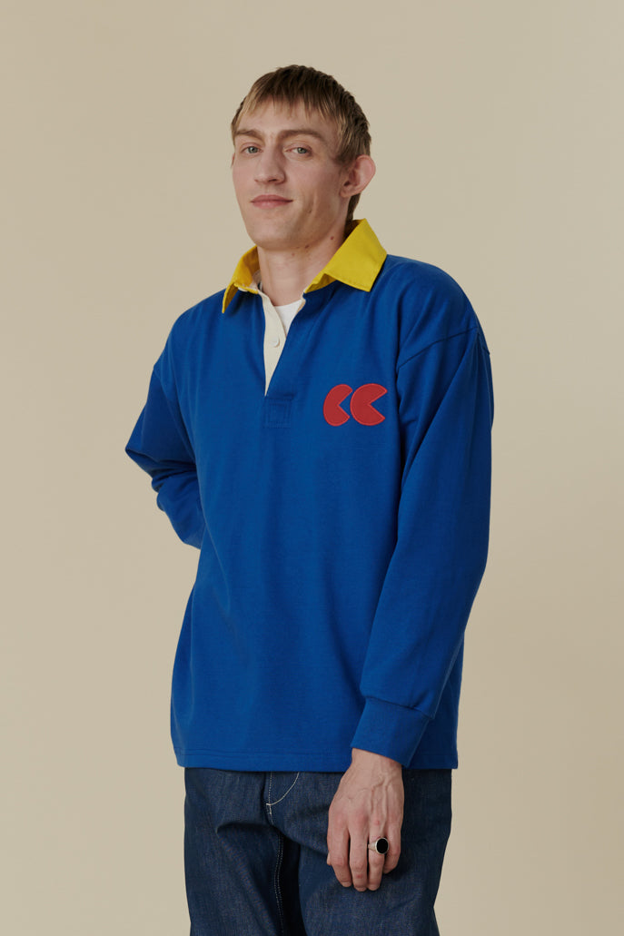 White male wearing long sleeve blue rugby shirt with contrast yellow collar and red CC logo badge on chest, worn over plain white t shirt.