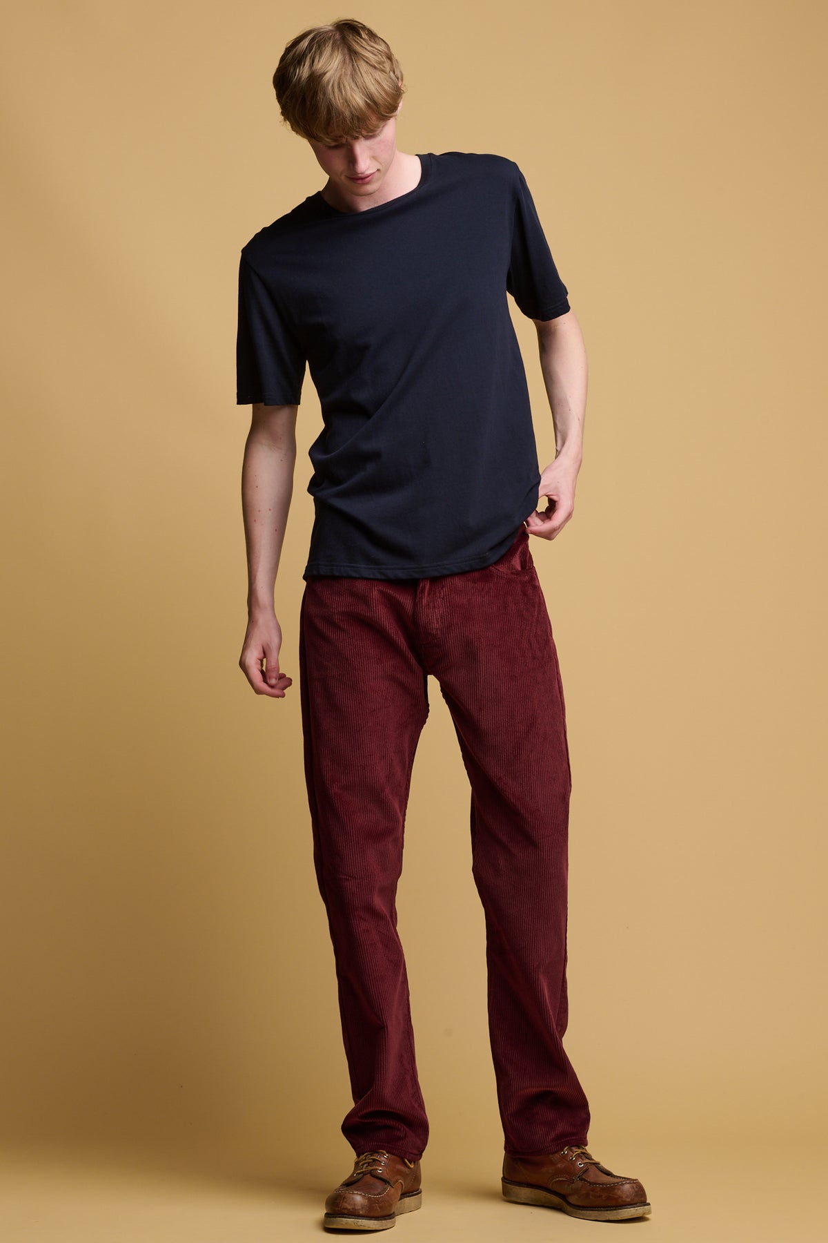 Male with blonde hair wearing community clothing five pocket cord trousers in burgundy paired with navy crew neck t shirt
