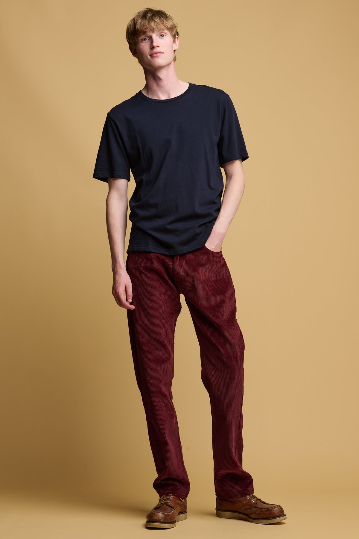 White male with blond hair wearing community clothing cord trousers in burgundy with his hand in one of the front pockets paired with short sleeve navy crew neck t shirt