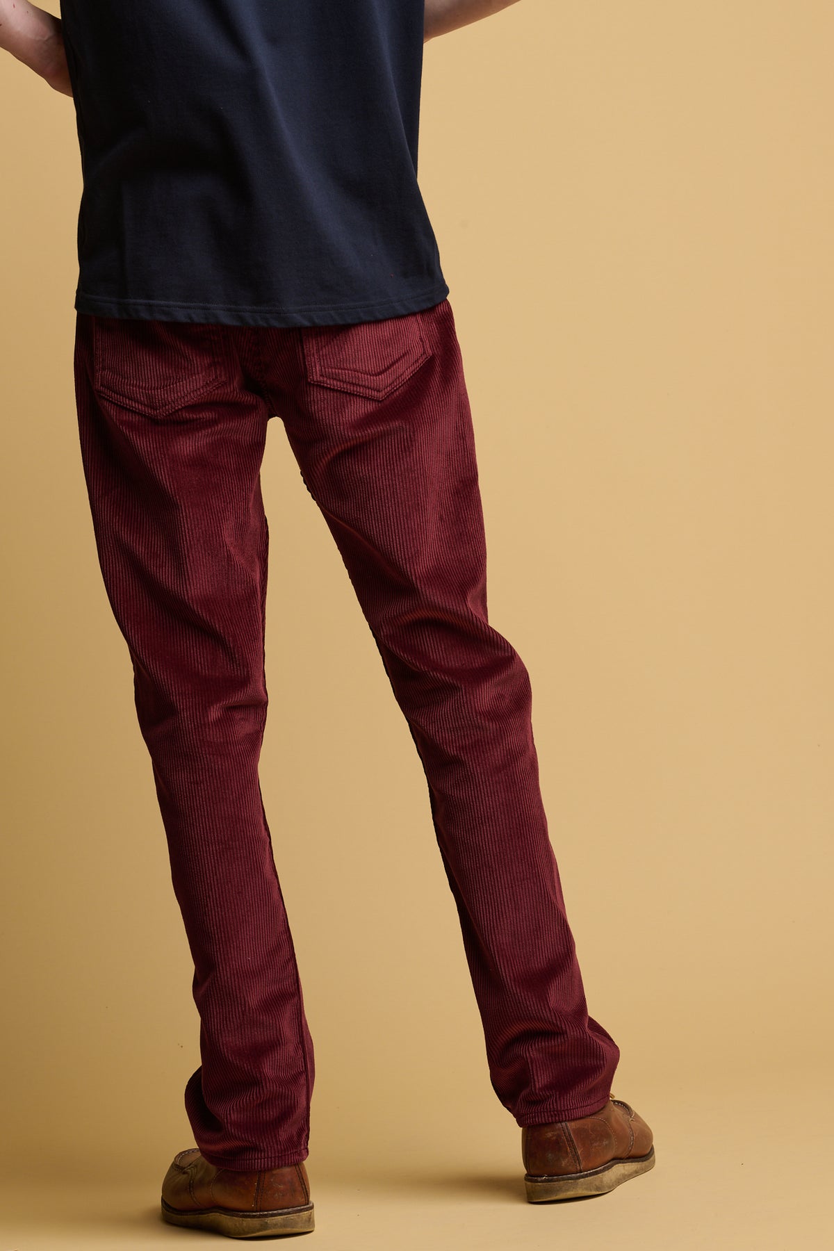 Shoulder down of the back of male wearing burgundy five pocket cord trousers with two back pockets paired with brown leather shoes and navy crew neck t shirt