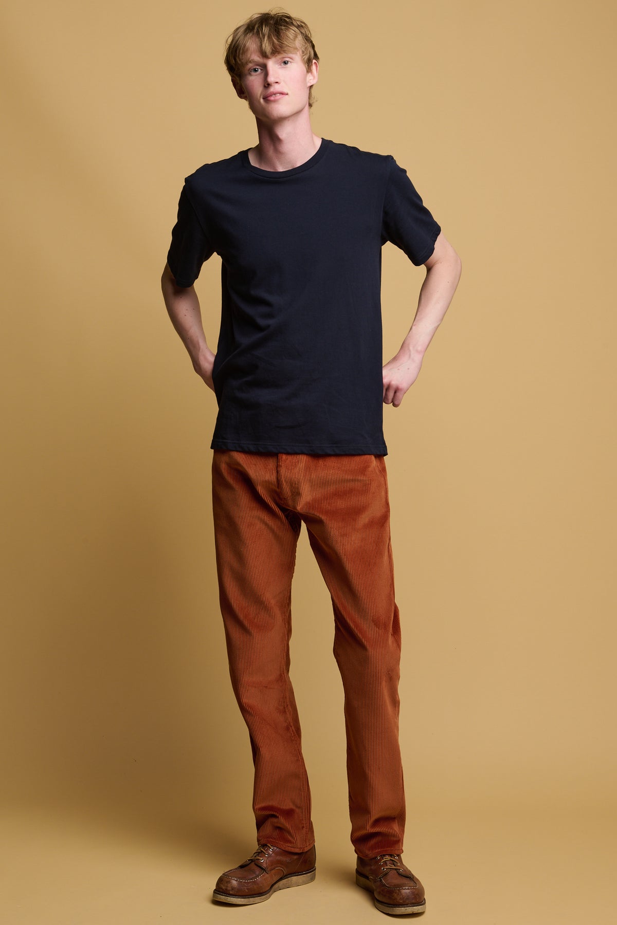 White male with dirty blonde hair holding his hands behind his hips wearing five pocket cord trousers in cinnamon paired with a short sleeve navy t shirt