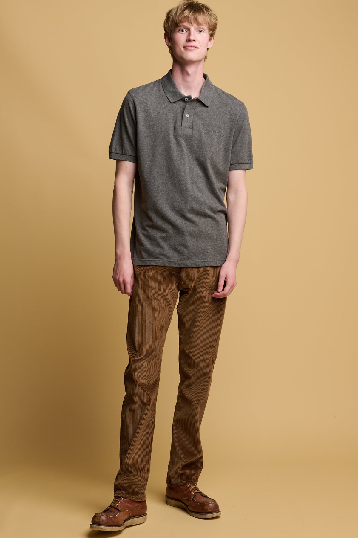 White male with dark blond hair wearing community clothing five pocket cord trousers in tan paired with short sleeve polo shirt in olive and brown leather shoes