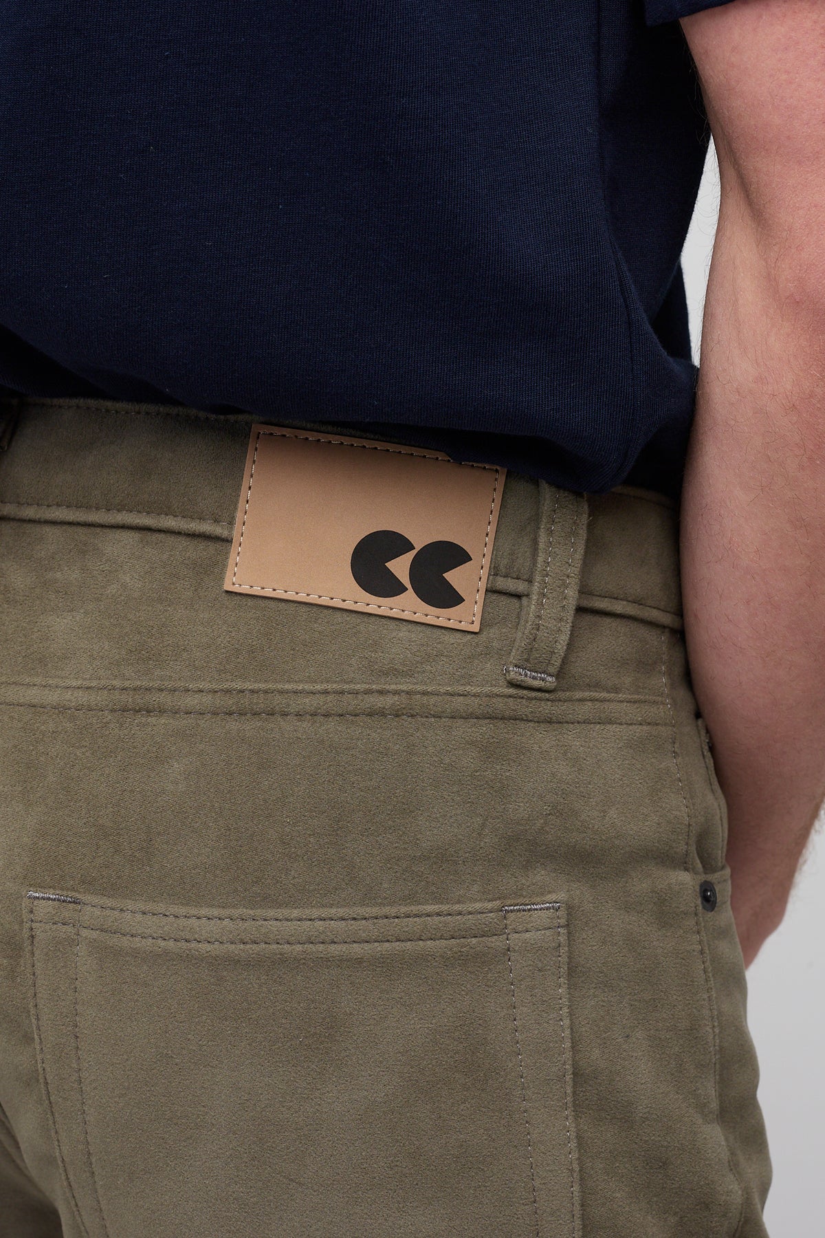 Brunette male in Olive Five Pocket Moleskin Jean styled with navy men's short sleeve t-shirt, brown CC logo badge close up