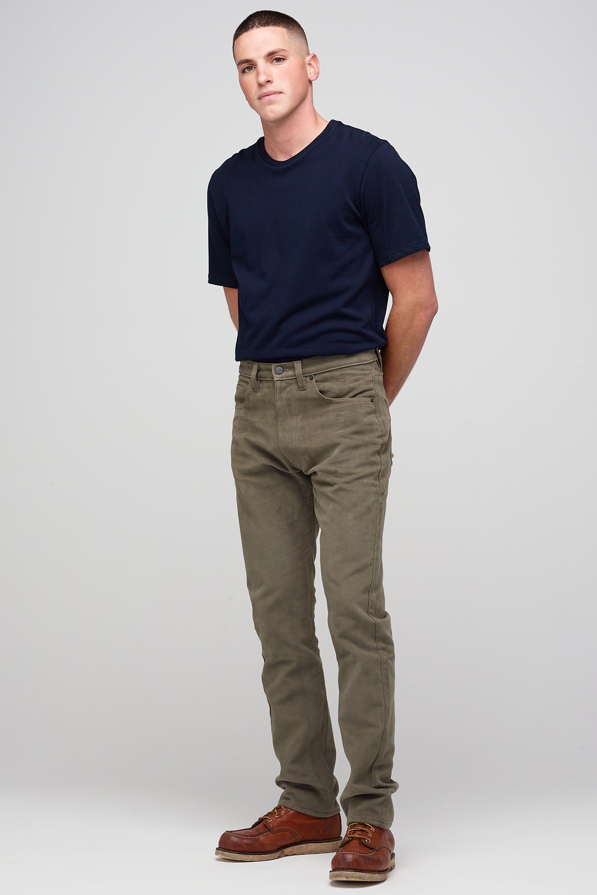 Brunette male in Olive Five Pocket Moleskin Jean styled with navy men's short sleeve t-shirt and brown leather shoes