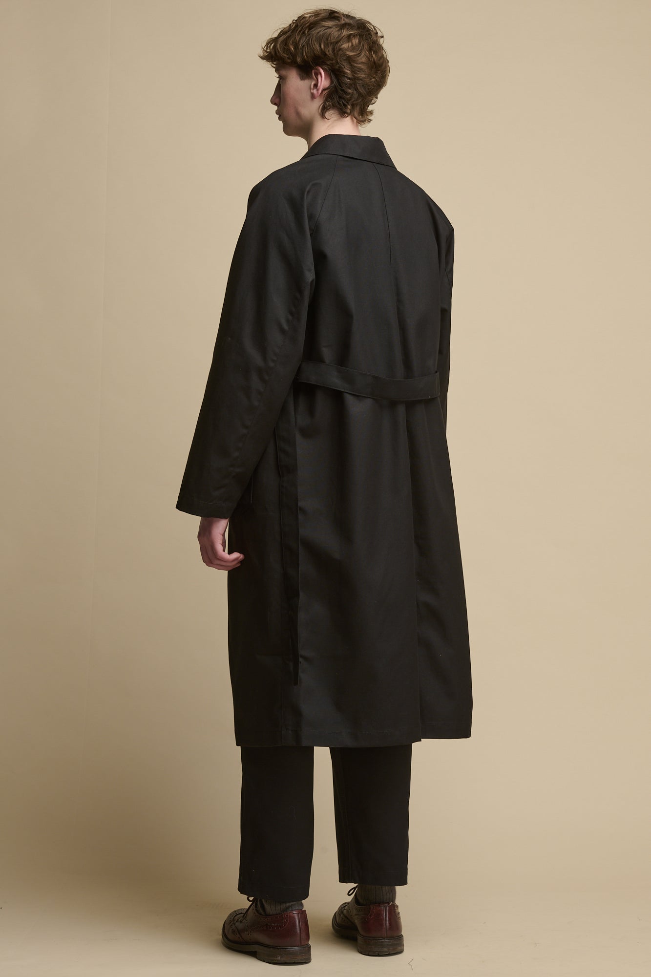 the back of male wearing Men's Frank Raglan Belted Raincoat in black. Belt held unfastened with two back belt loops 