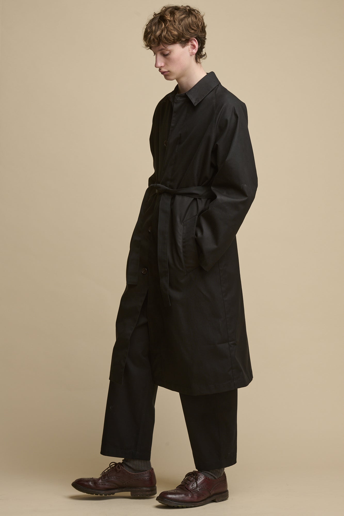 Full body image of white male wearing fastened Men's Frank Raglan Belted Raincoat in black with his hands in the two front pockets