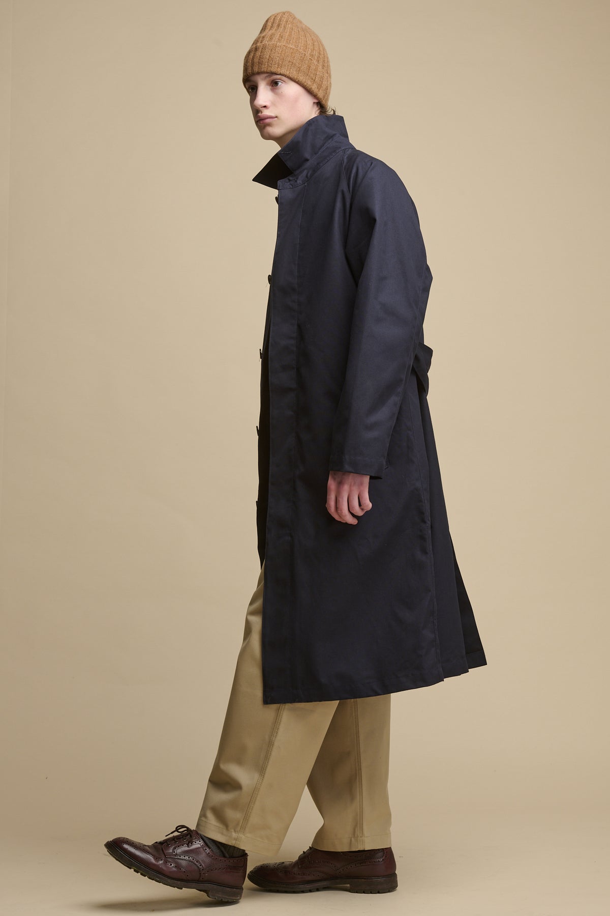 the side of male wearing Frank Raglan Belted Raincoat in navy with a dark camel beanie worn with men's cameraman pants in putty