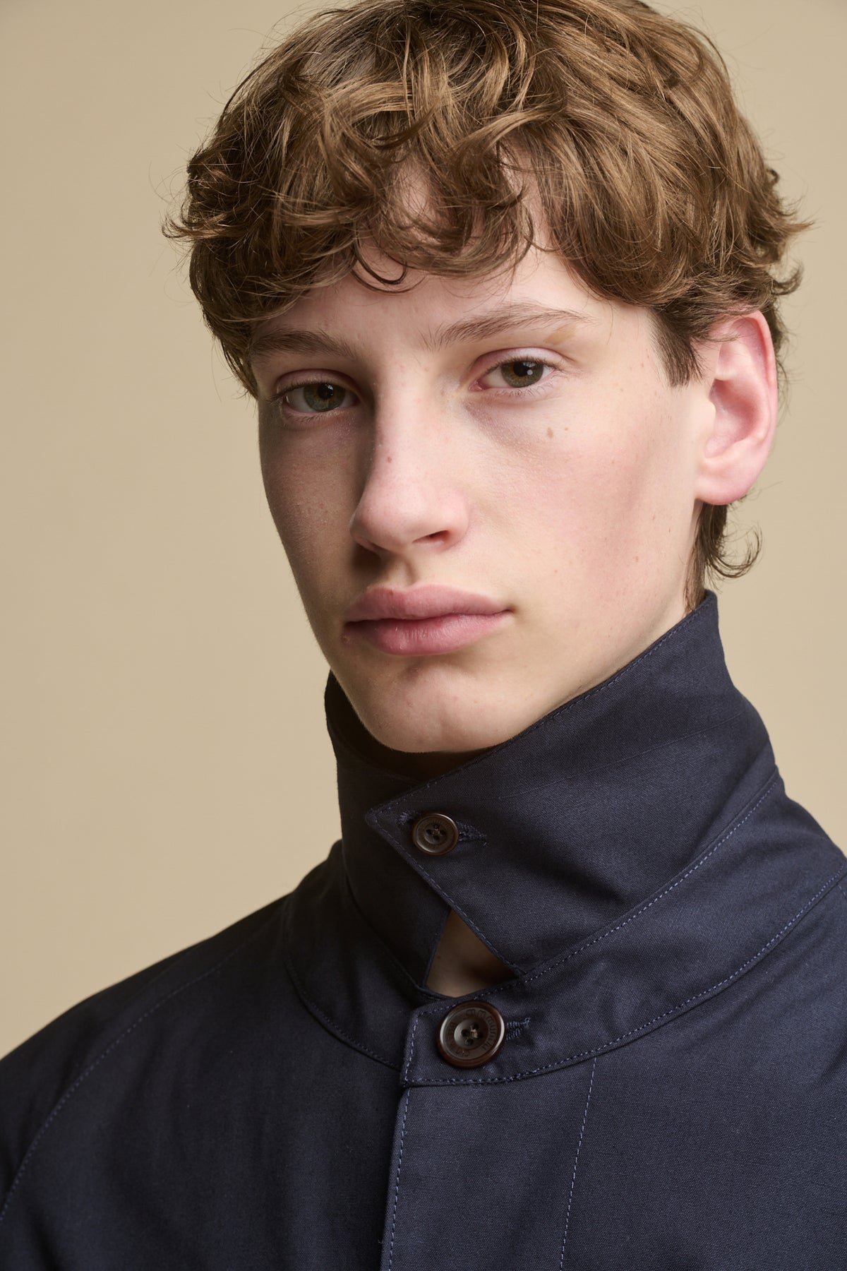 Portrait of male wearing collar up of the Men's Frank Raglan Belted Raincoat in navy with community clothing logo brown buttons