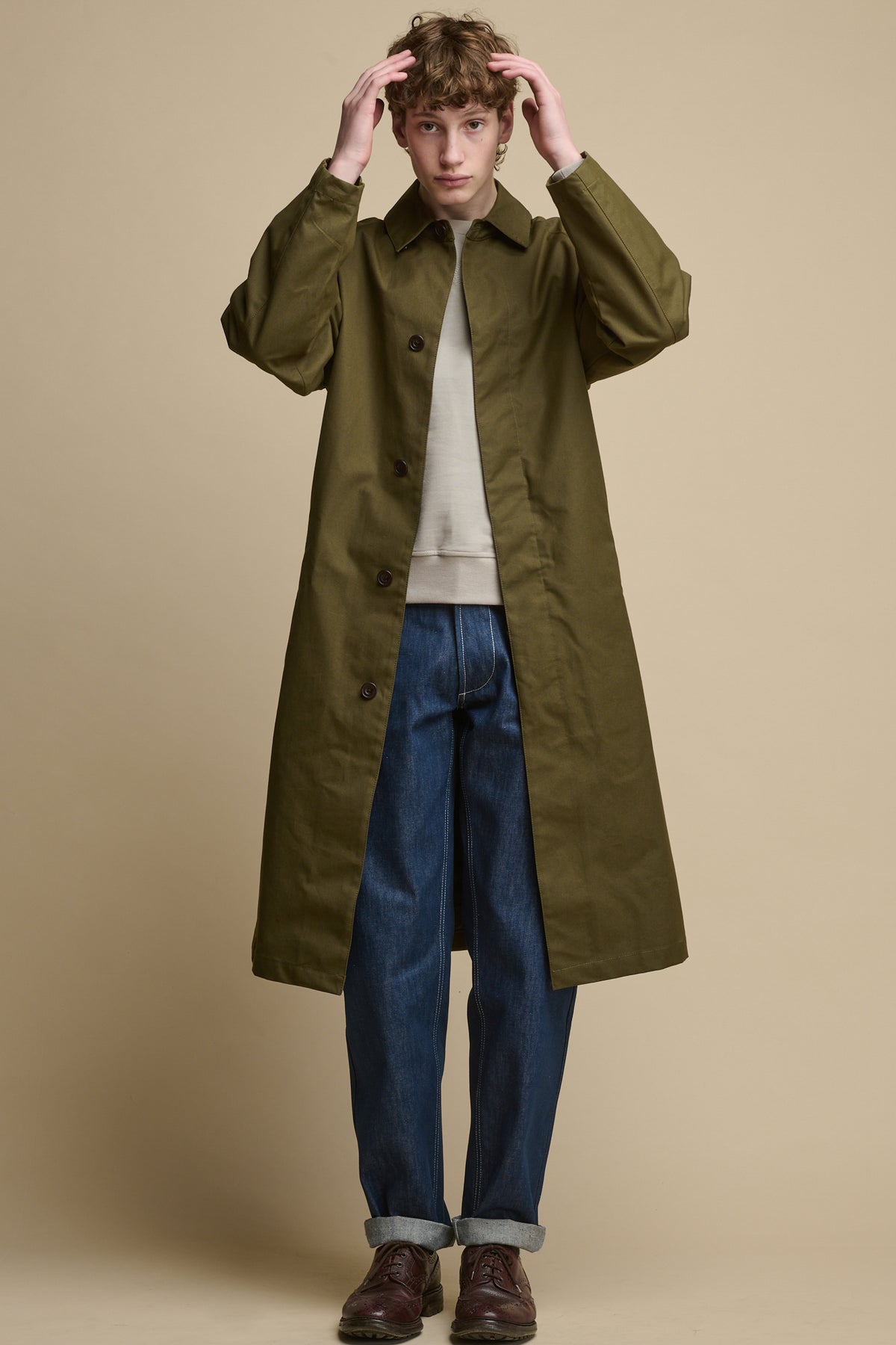 Full body image of the front of white male wearing Frank Raglan Belted Raincoat in Olive, unfastened worn over raglan sweatshirt in bone with chore jeans in blue and brown leather shoes