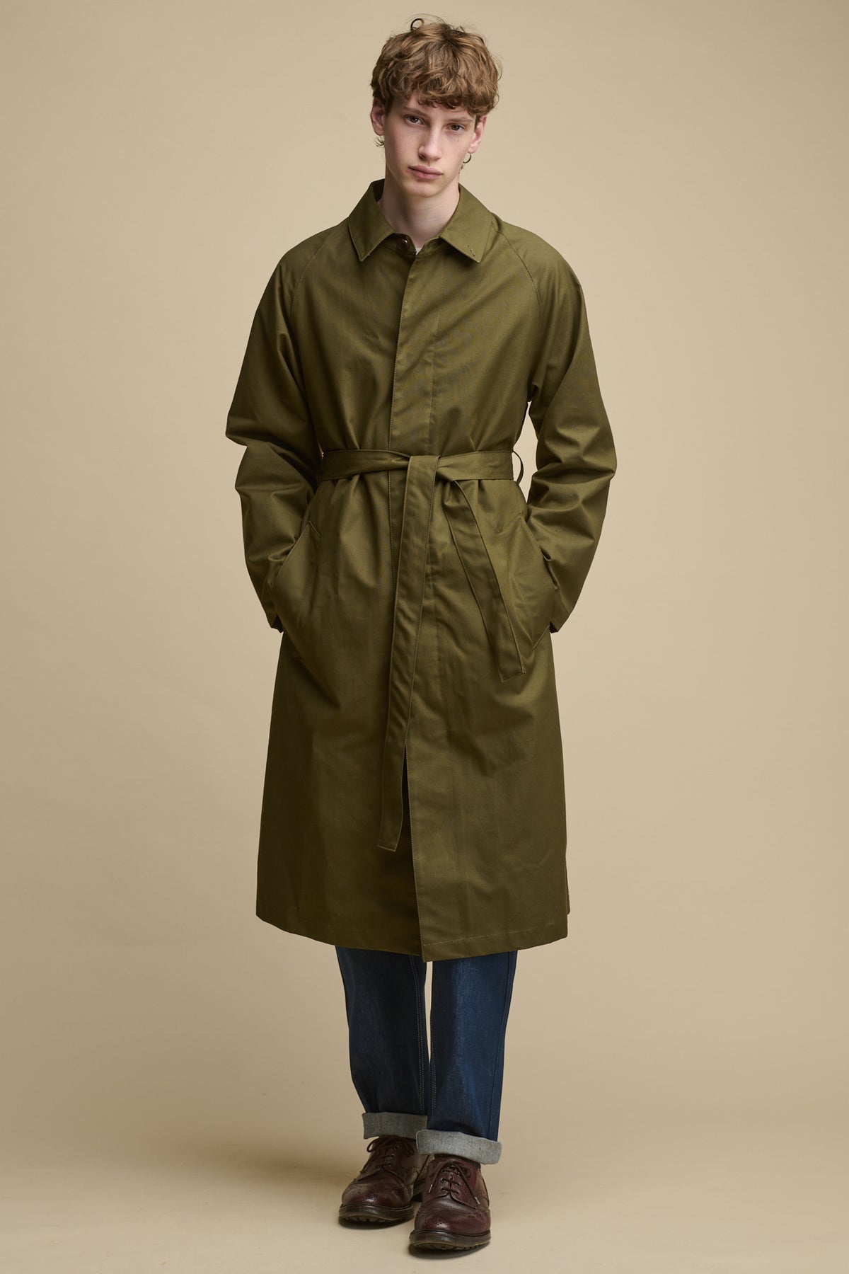 Full body image of the front of white mail wearing fastened Frank Raglan Belted Raincoat in Olive with his hands in the front two pockets