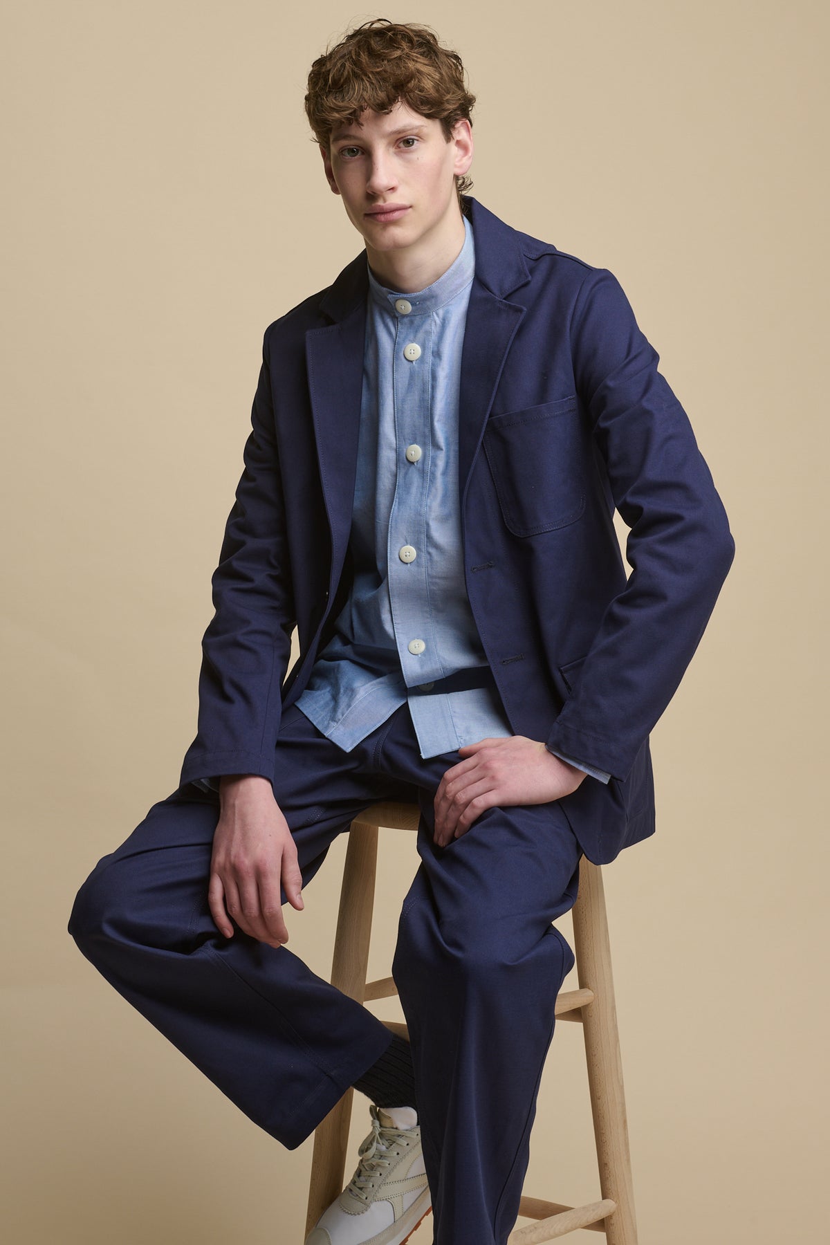 
            Full body image of male sat on stool wearing George Lightweight Collarless Overshirt in light blue paired with James Single Breasted Blazer in navy with cameraman pants in Navy