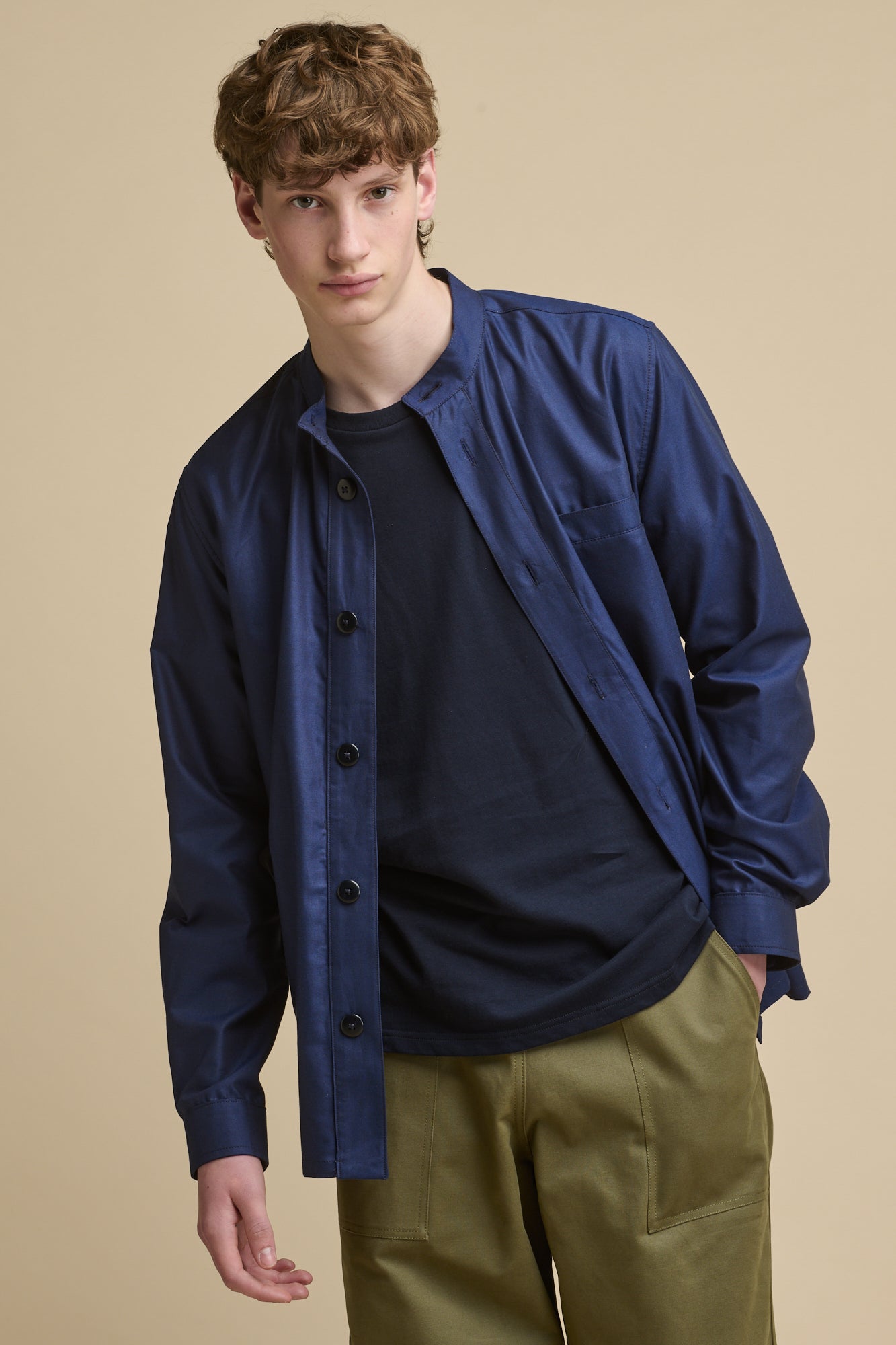 Thigh up image of male wearing George Lightweight Collarless Overshirt unbuttoned over navy t shirt paired with Cameraman Pants in olive