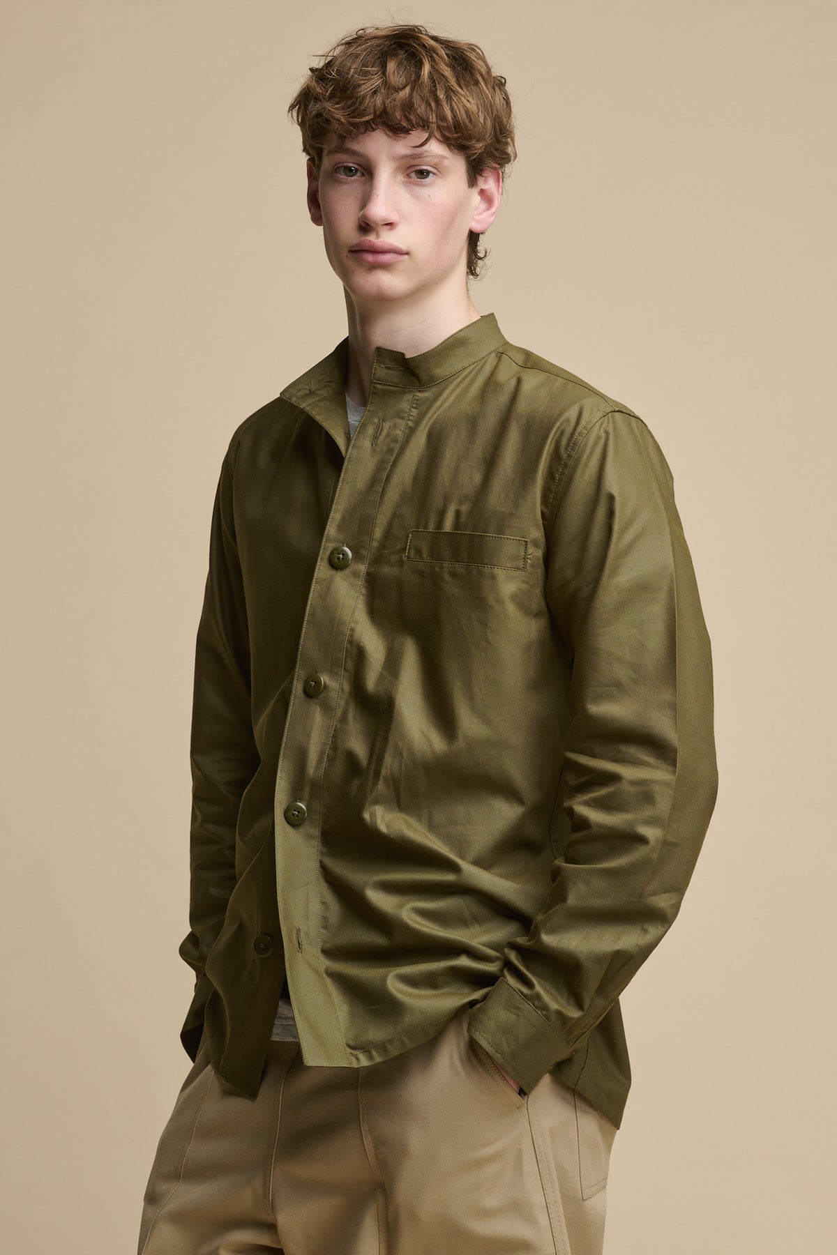 
            Male wearing men&#39;s George Lightweight Collarless Overshirt in Olive worn with half unbuttoned over grey t shirt paired with Cameraman pants in putty 