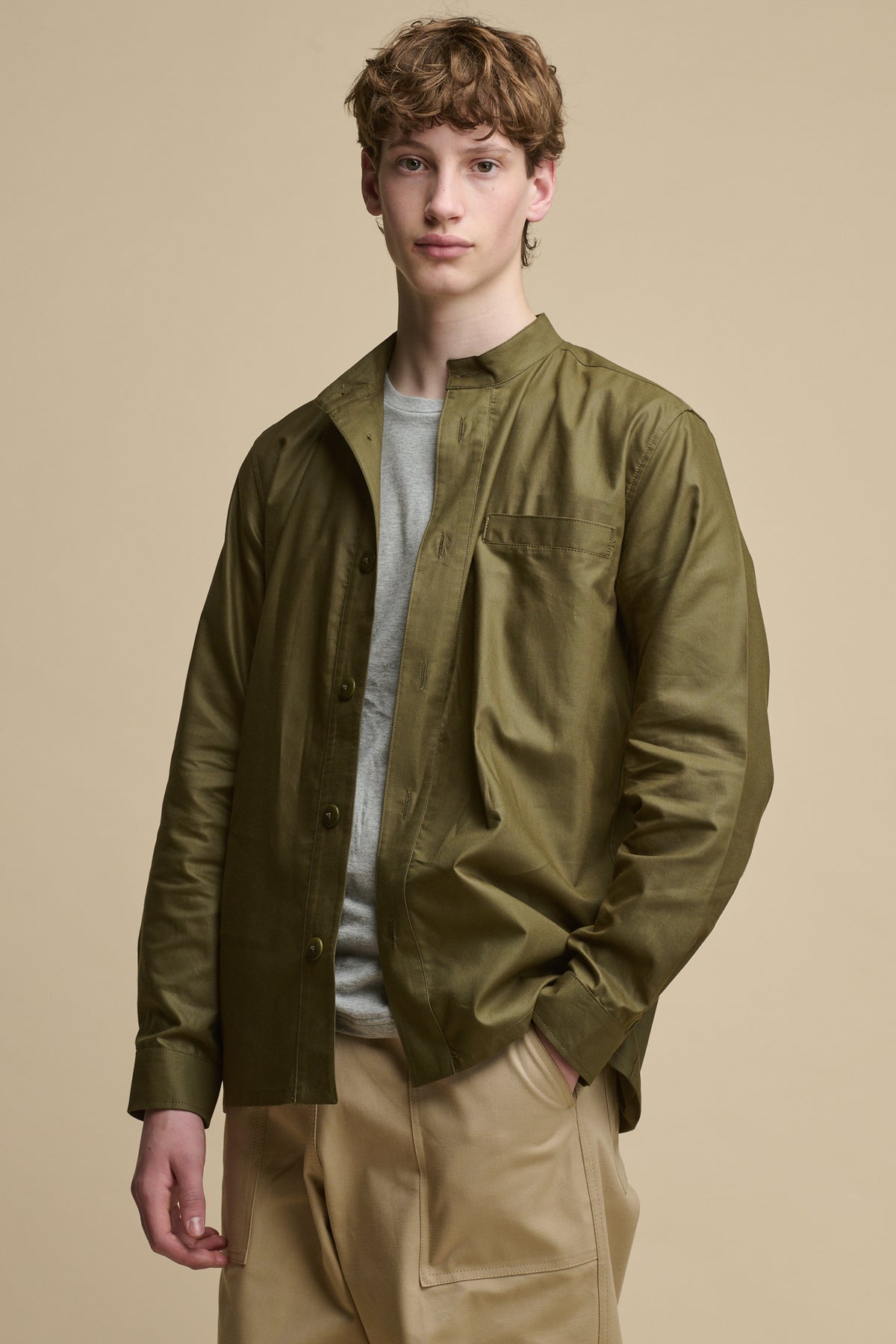 
            White male wearing unbuttoned men&#39;s George Lightweight Collarless Overshirt in Olive worn over grey t shirt paired with Cameraman pants in putty 