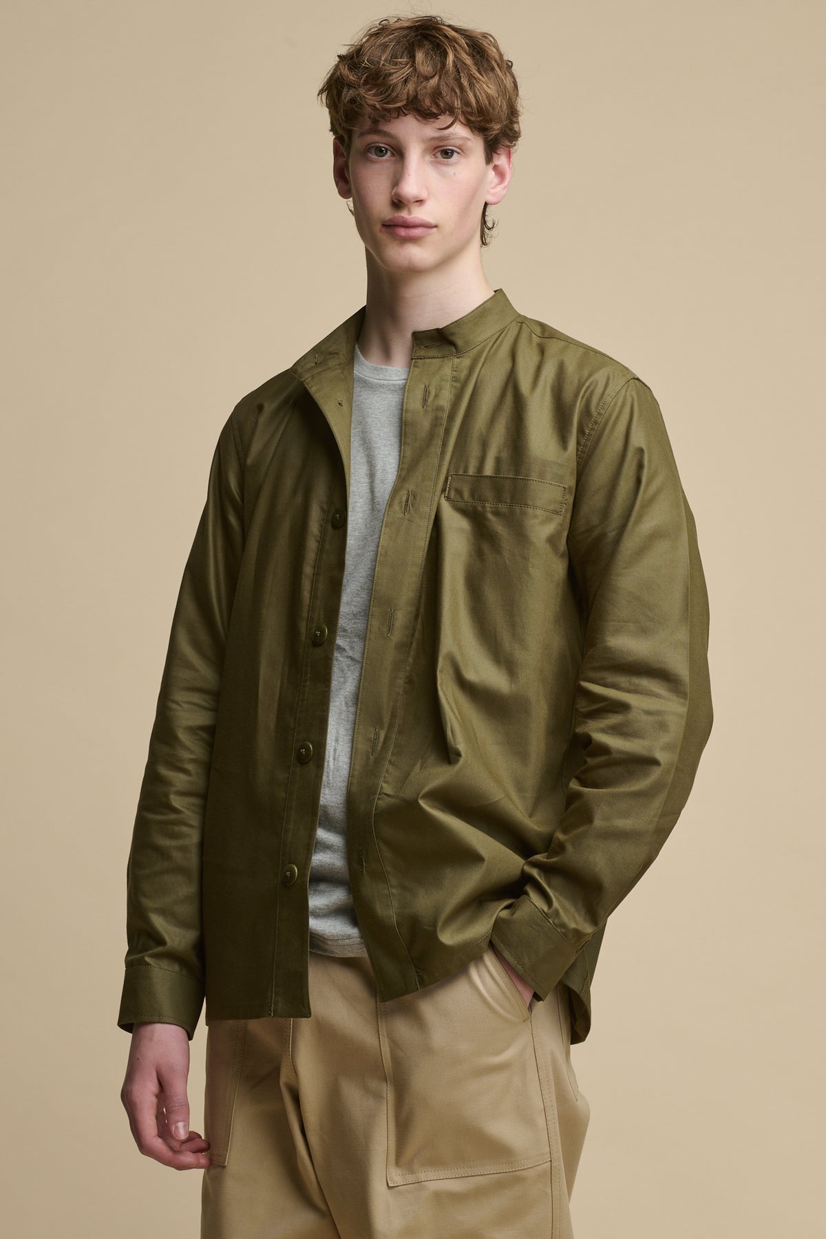 White male wearing unbuttoned men's George Lightweight Collarless Overshirt in Olive worn over grey t shirt paired with Cameraman pants in putty