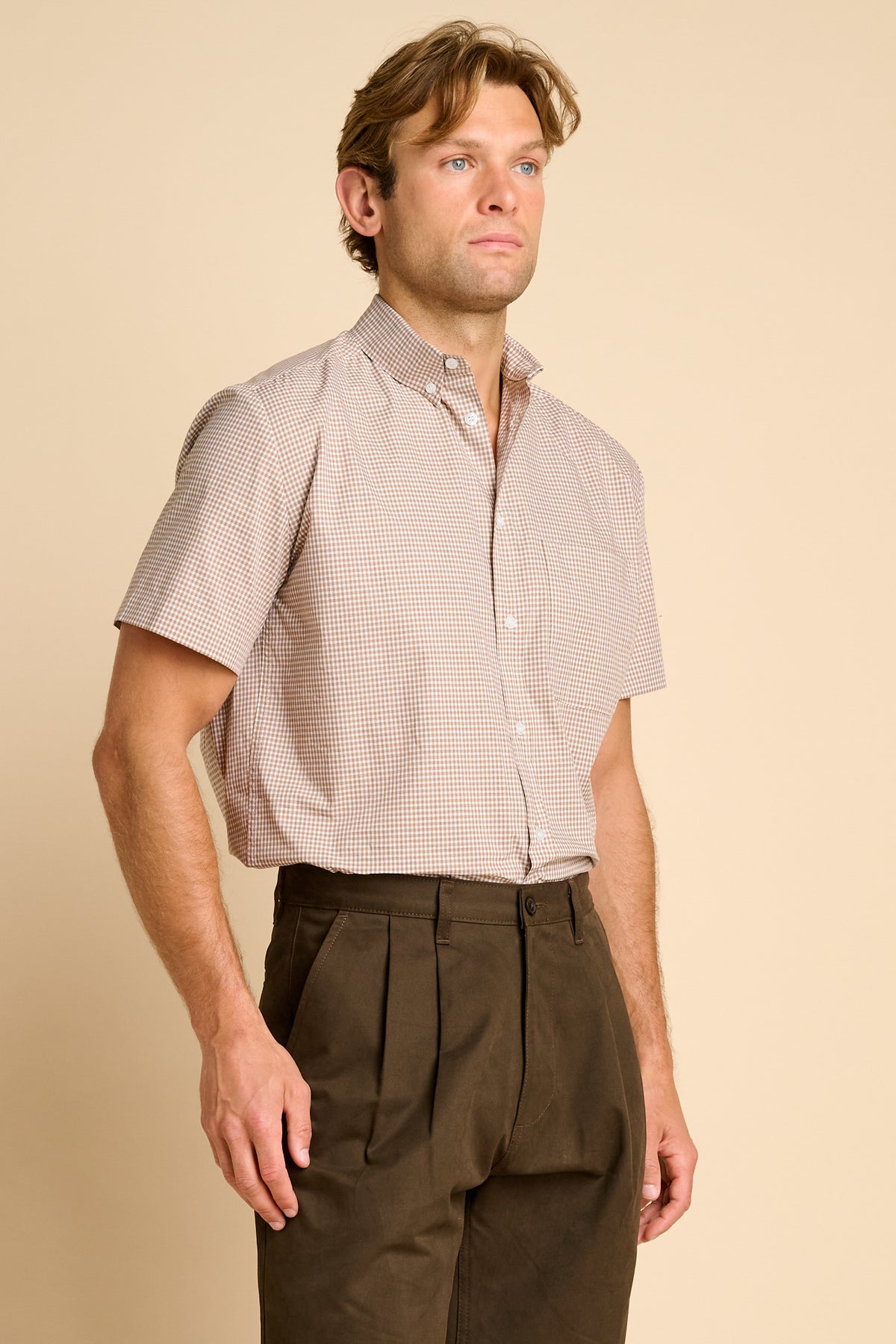 A man wearing a short sleeve button down shirt paired with dark brown pleated trousers, against a beige background.