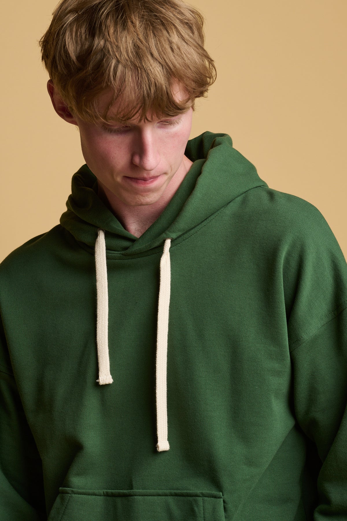 close up of male looking down wearing hooded sweatshirt in bottle green with ecru drawcord through the hood