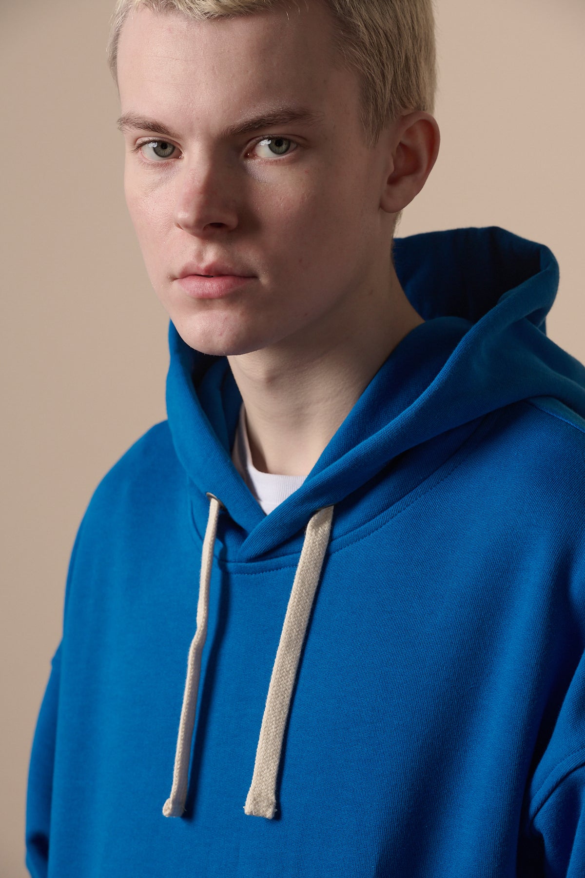 Portrait of blonde male wearing cobalt hooded sweatshirt with ecru drawcord through the hood. White t shirt underneath.