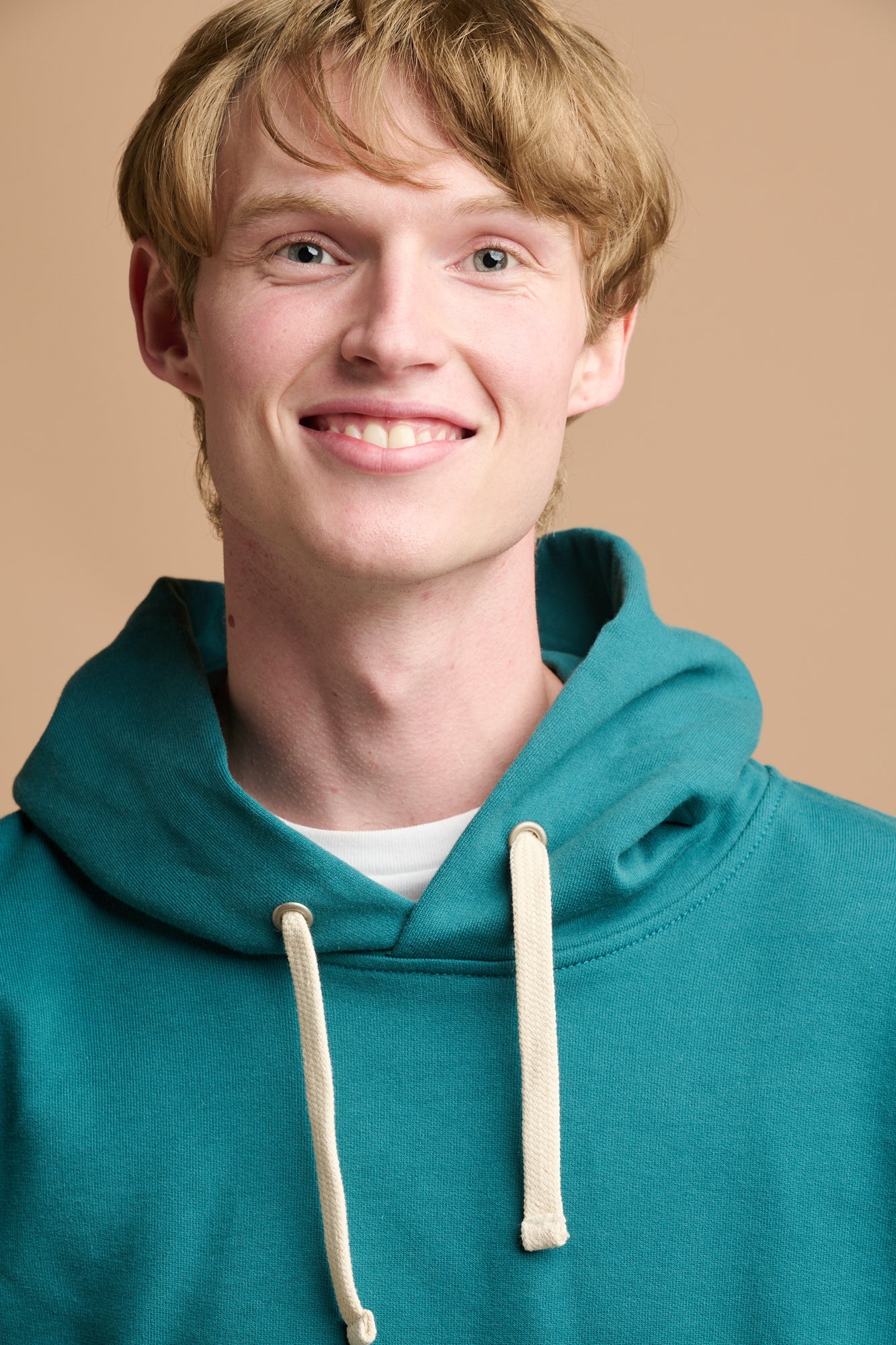 Close up head shot of smiley male with blonde hair wearing hooded sweatshirt in teal over white crew neck t shirt