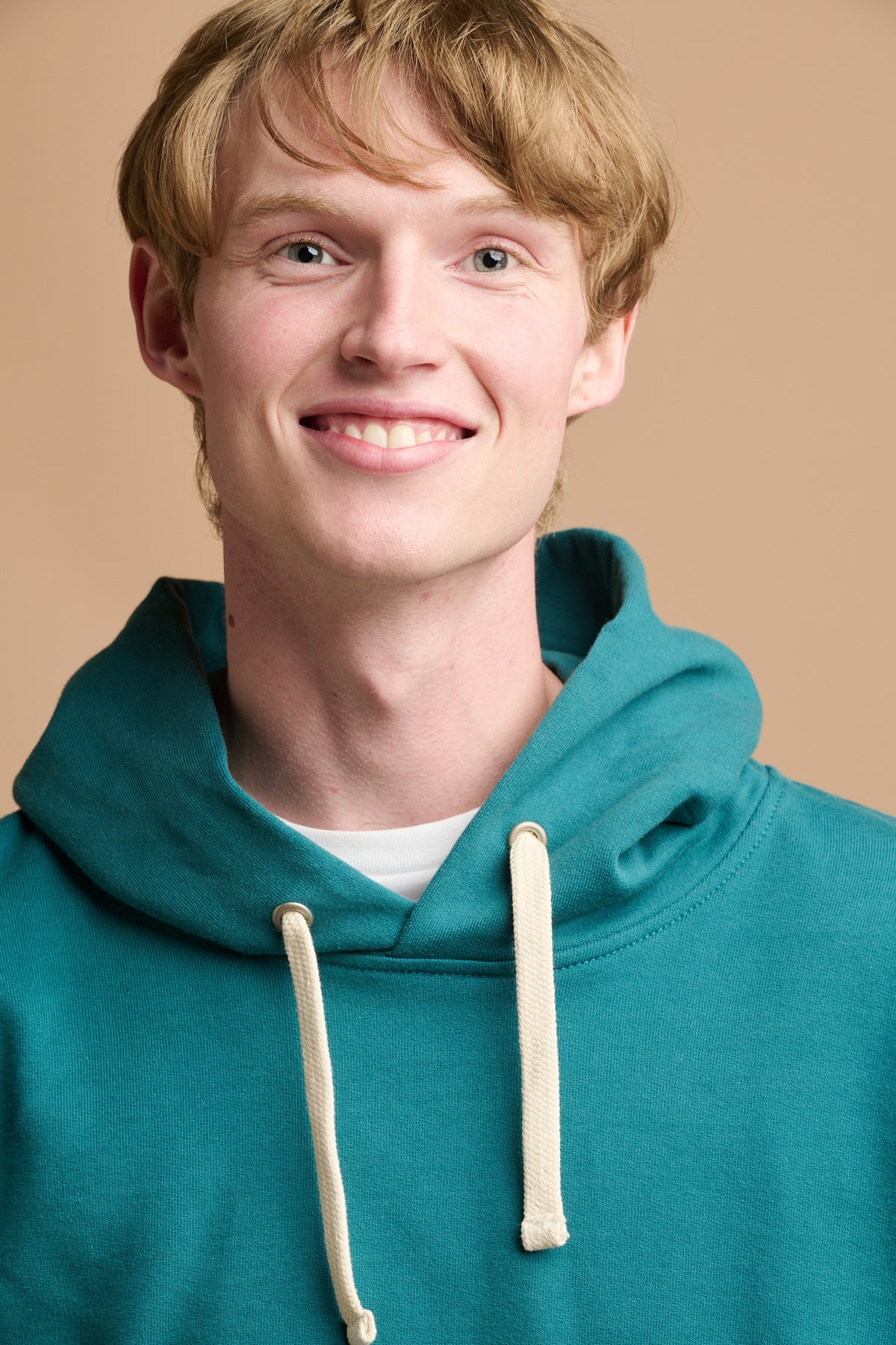 Close up head shot of smiley male with blonde hair wearing hooded sweatshirt in teal over white crew neck t shirt