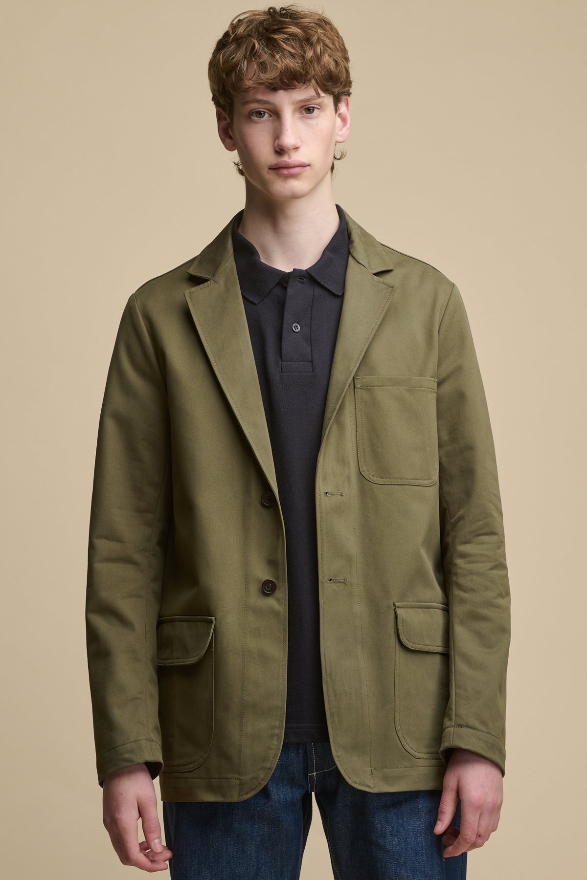 Thigh up image of male wearing Men's James Single Breasted Blazer in olive worn over men's short sleeve polo shirt in navy with blue jeans. Blazer has two button fastening, two front pocket with flaps and one chest pocket