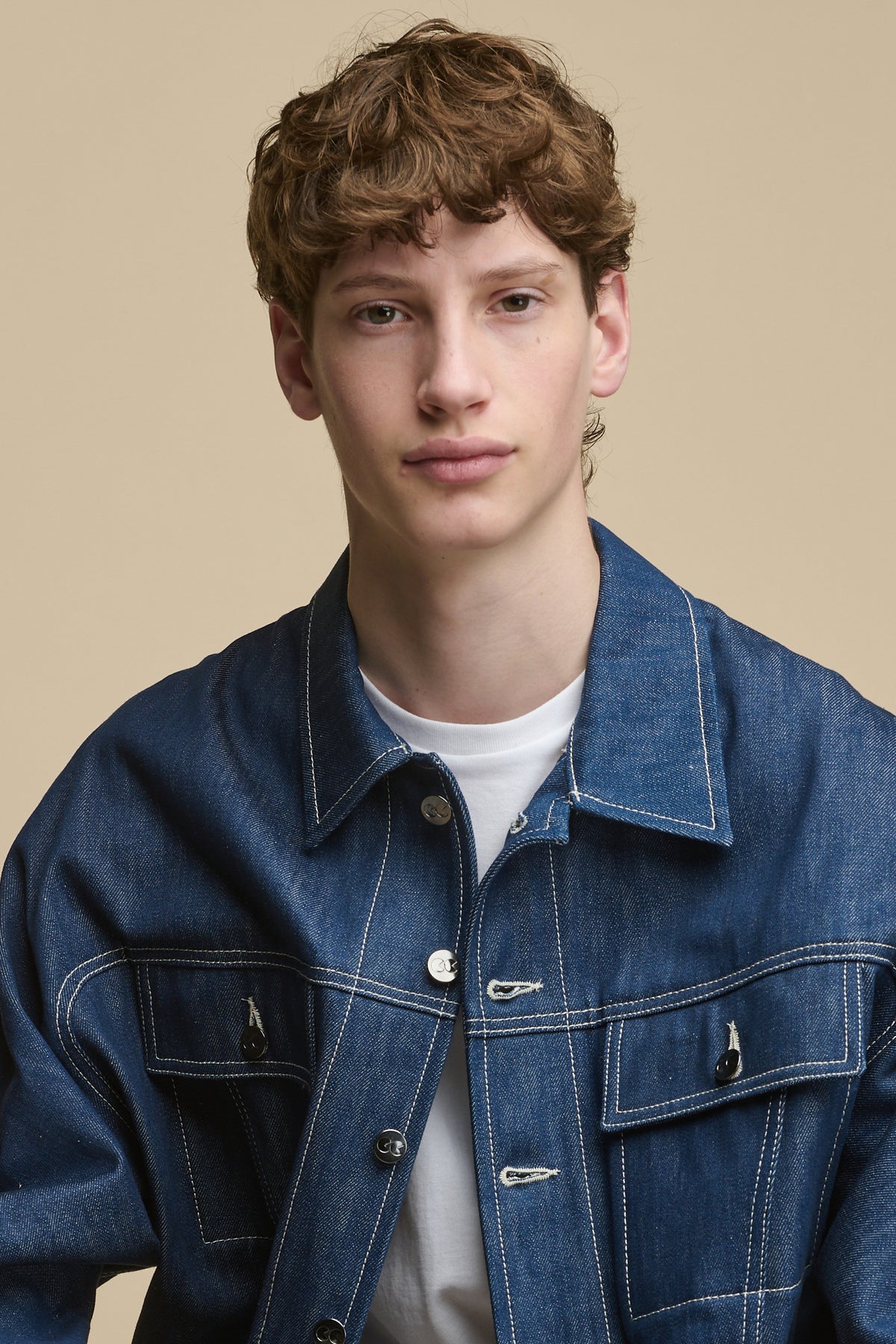 Portrait of white male wearing Joe Denim Jacket in blue over short sleeve t shirt in white. Jacket has contrast white stitching and silver branded buttons, two front pockets with flaps