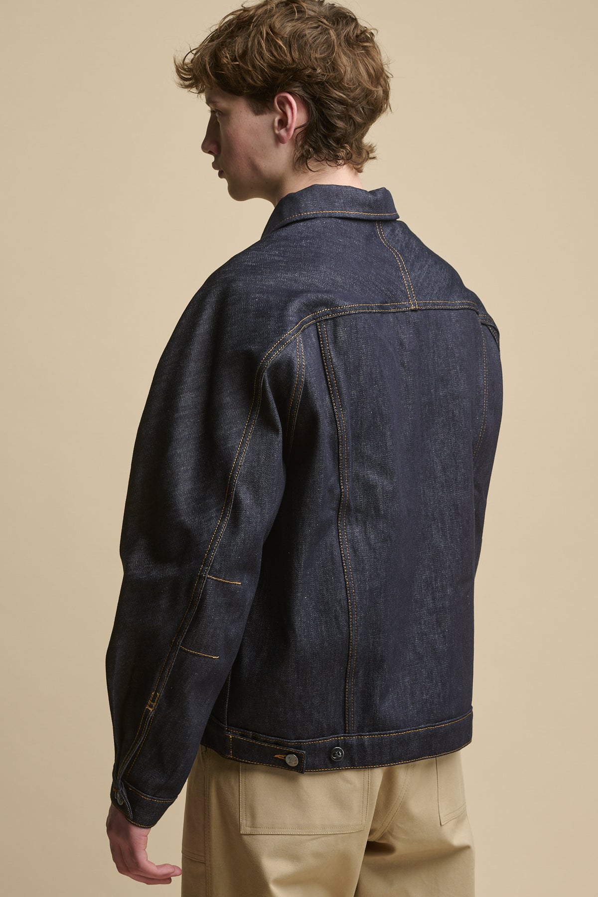 Thigh up image of the back of male wearing Joe Denim Jacket in indigo, adjustable button fastening detail on hem