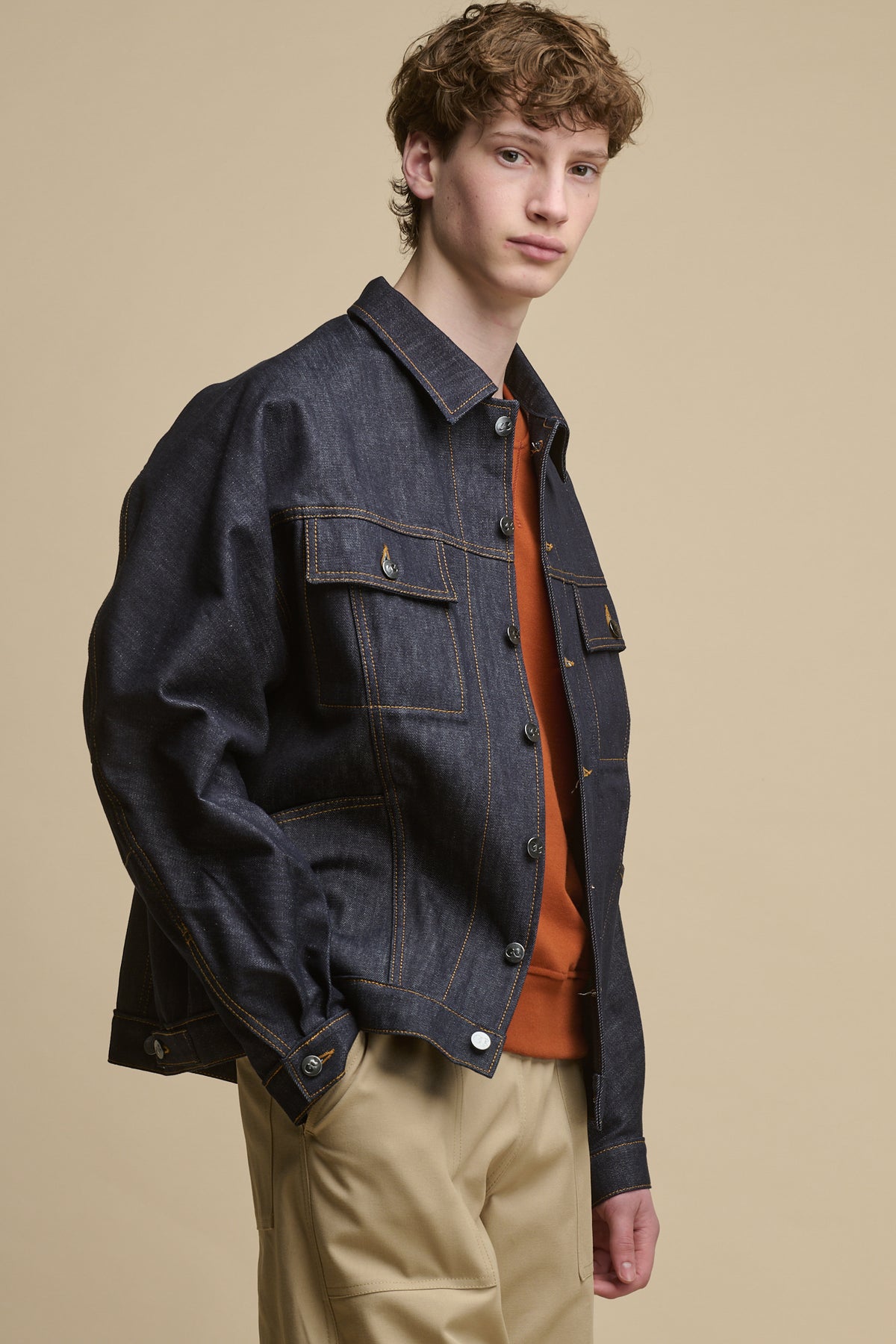 Thigh up image of male standing on an angle looking towards the camera wearing Joe Denim Jacket in indigo worn with raglan sweatshirt in cinnamon and cameraman pants in putty