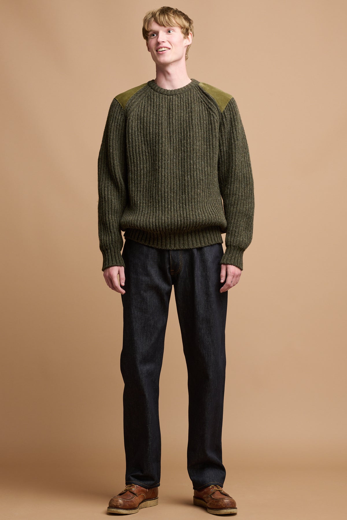 The front of male wearing lambswool fisherman ribbed jumper in dark olive paired with loose fit button fly arran jean in indigo