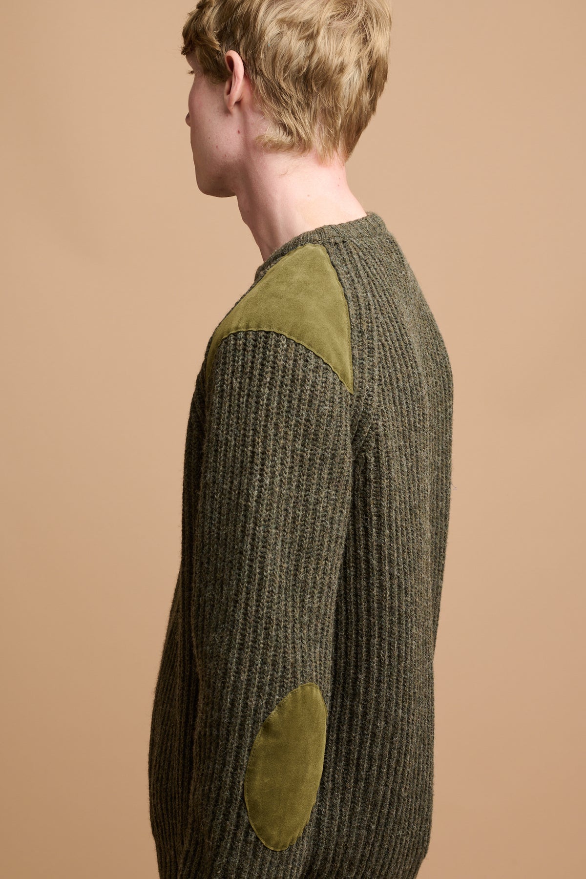 The side of male wearing lambswool fisherman ribbed jumper in dark olive with suede patches on the shoulder and elbow