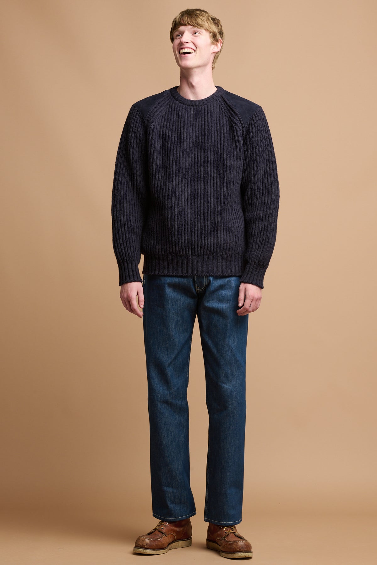 
            Smiley male wearing community clothing lambswool fisherman ribbed jumper in navy paired with loose fit button fly blue jeans