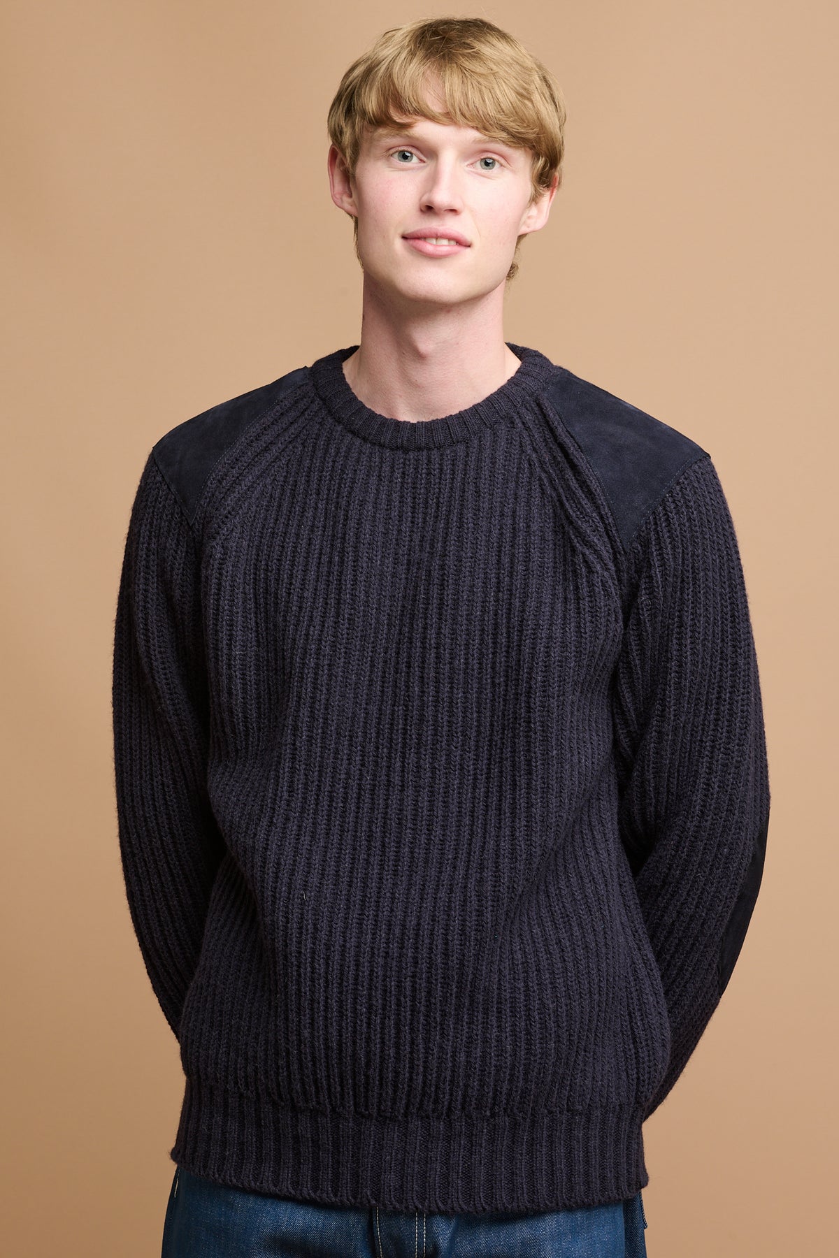 Hip up front of male with his hands held behind wearing lambswool fisherman ribbed jumper in navy