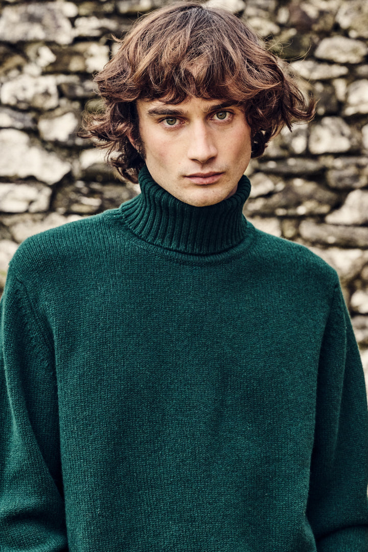 Man wearing a green roll neck jumper against a stone wall background
