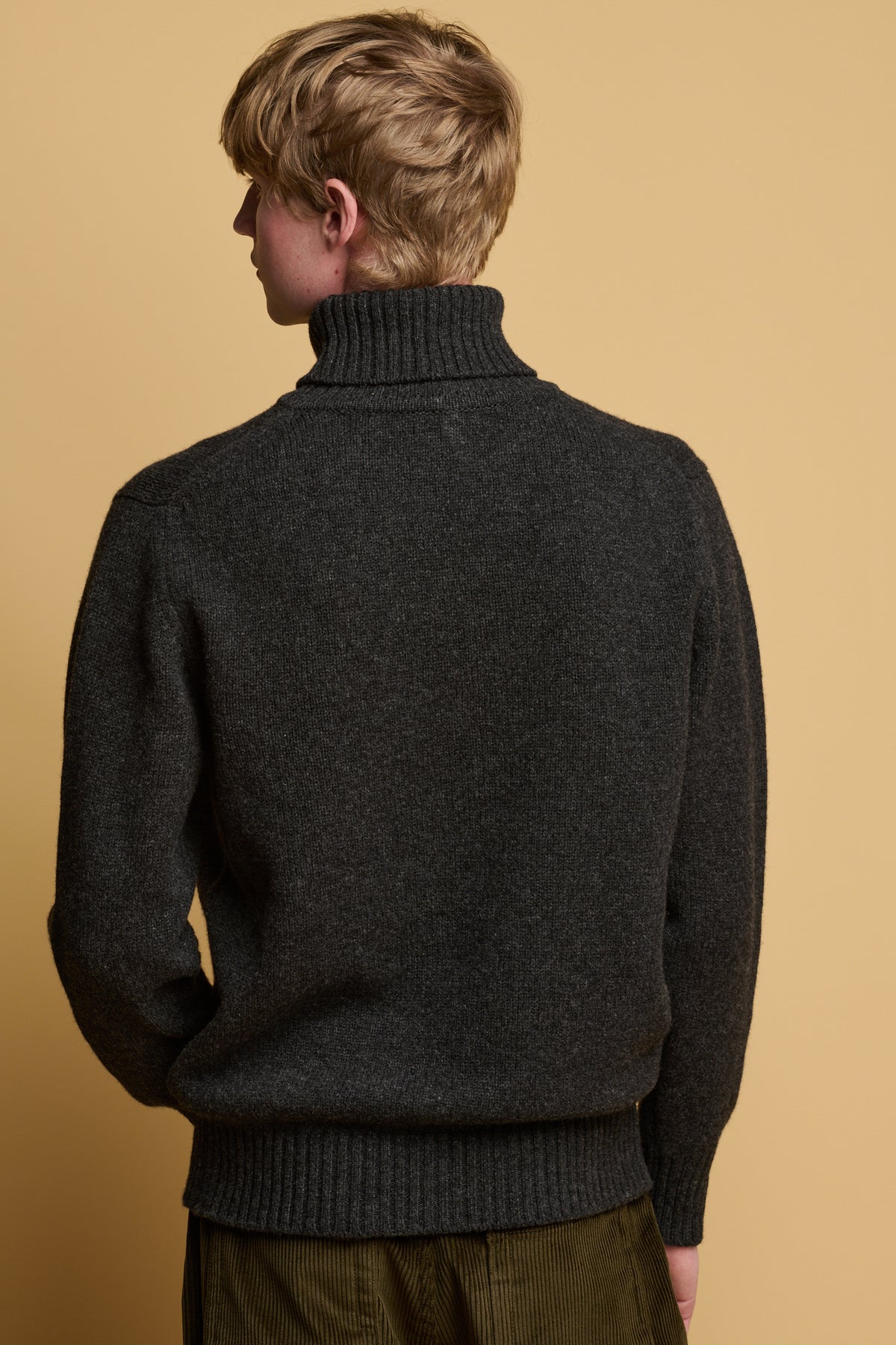 Thigh up image of the back of male wearing lambswool roll neck in charcoal paired with cord trousers in olive