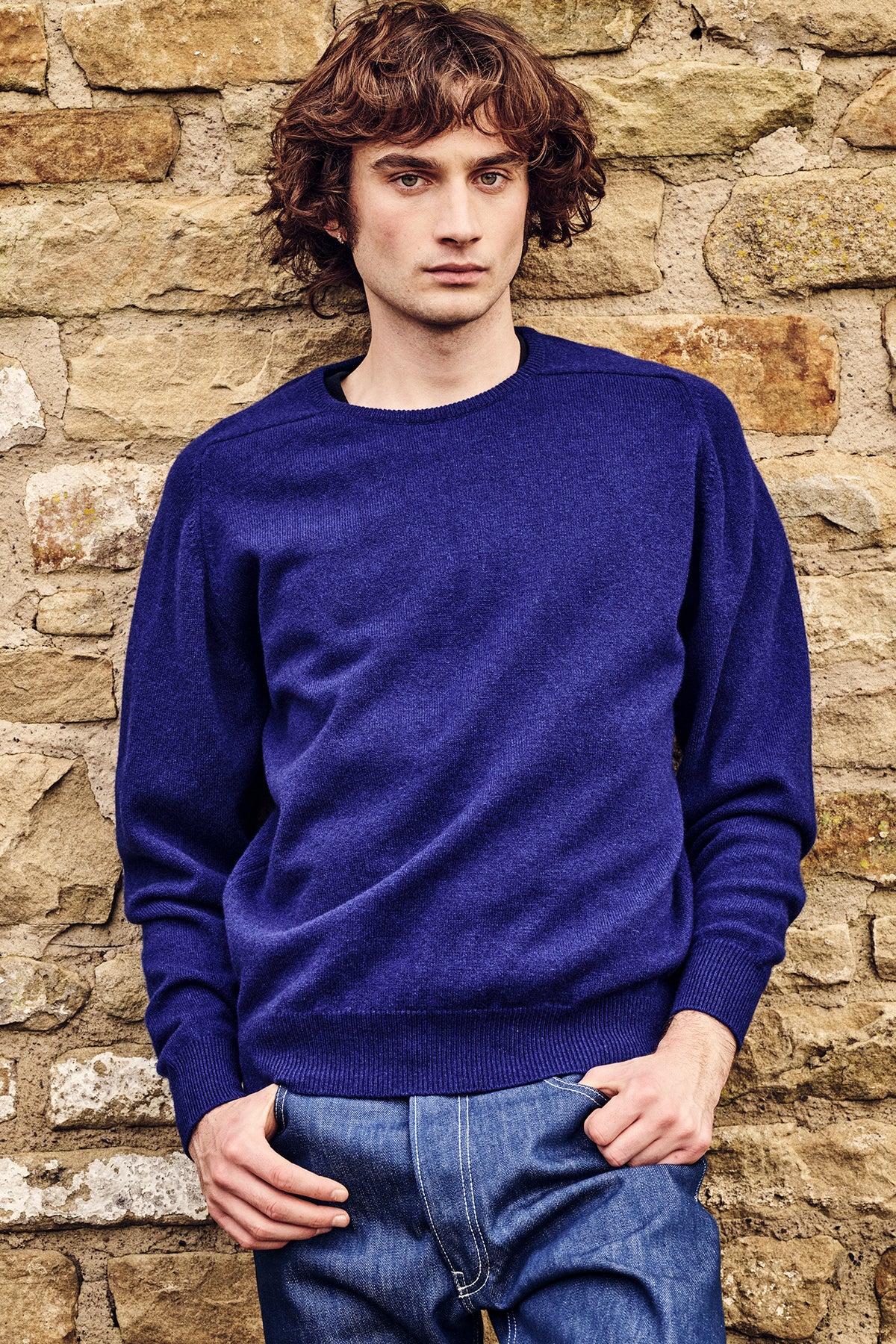 Man wearing a blue sweater and jeans standing against a stone wall.