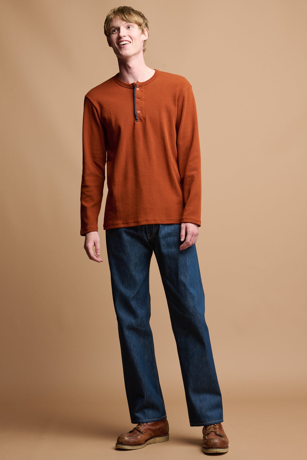 
            Smiley male wearing long sleeve henley top in cinnamon paired with loose button fly arran jean in blue