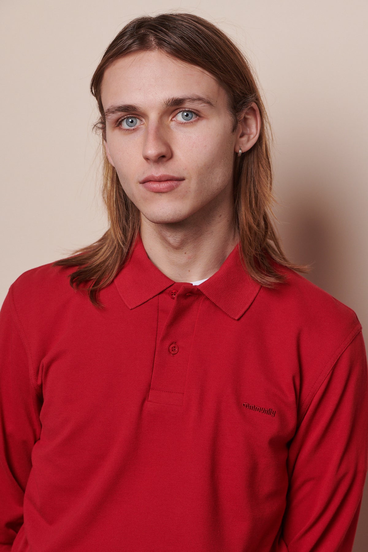 
            Portrait of white male with shoulder length hair wearing long sleeve polo shirt with top button undone. Community stitched into chest of shirt in matching crimson