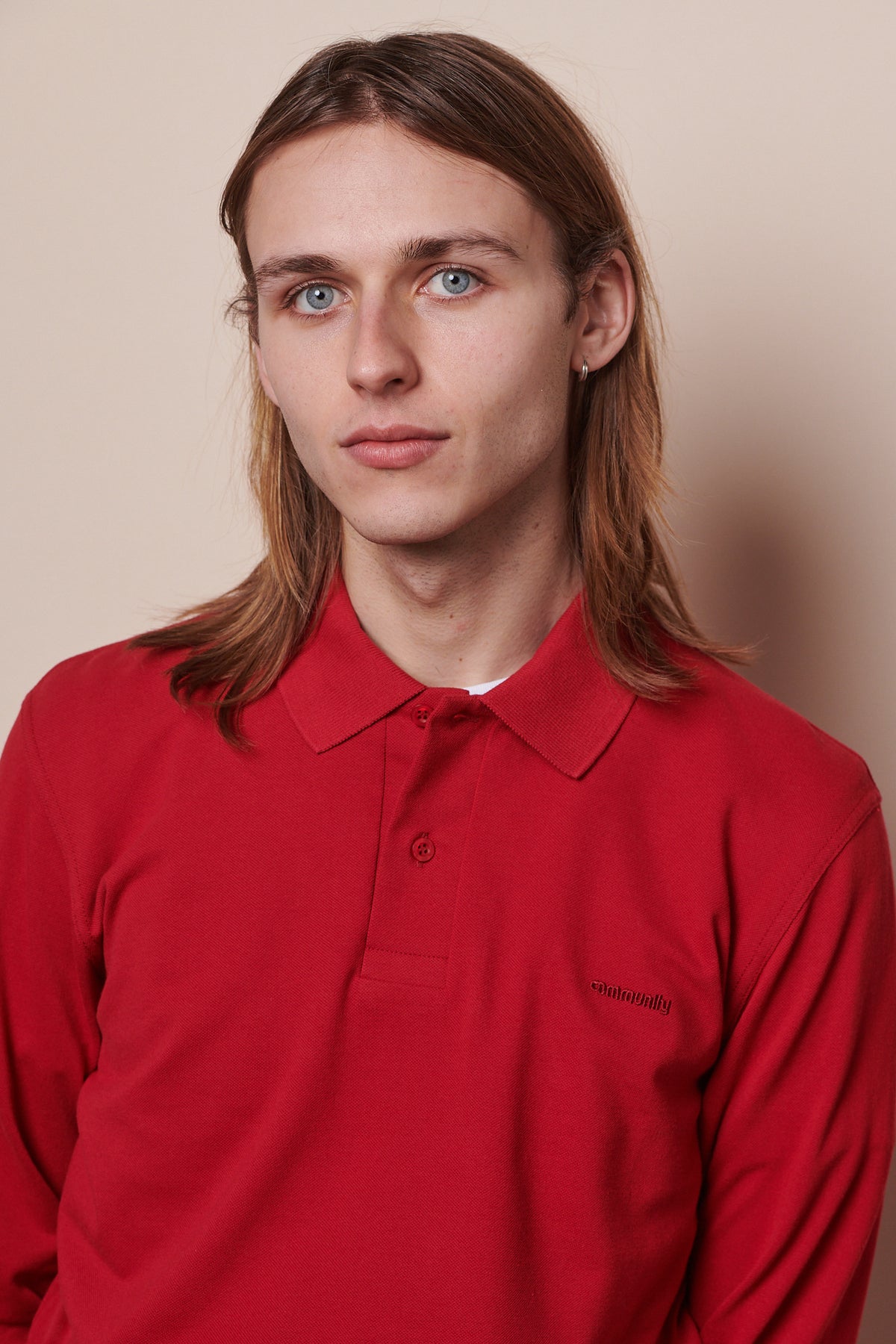 Portrait of white male with shoulder length hair wearing long sleeve polo shirt with top button undone. Community stitched into chest of shirt in matching crimson