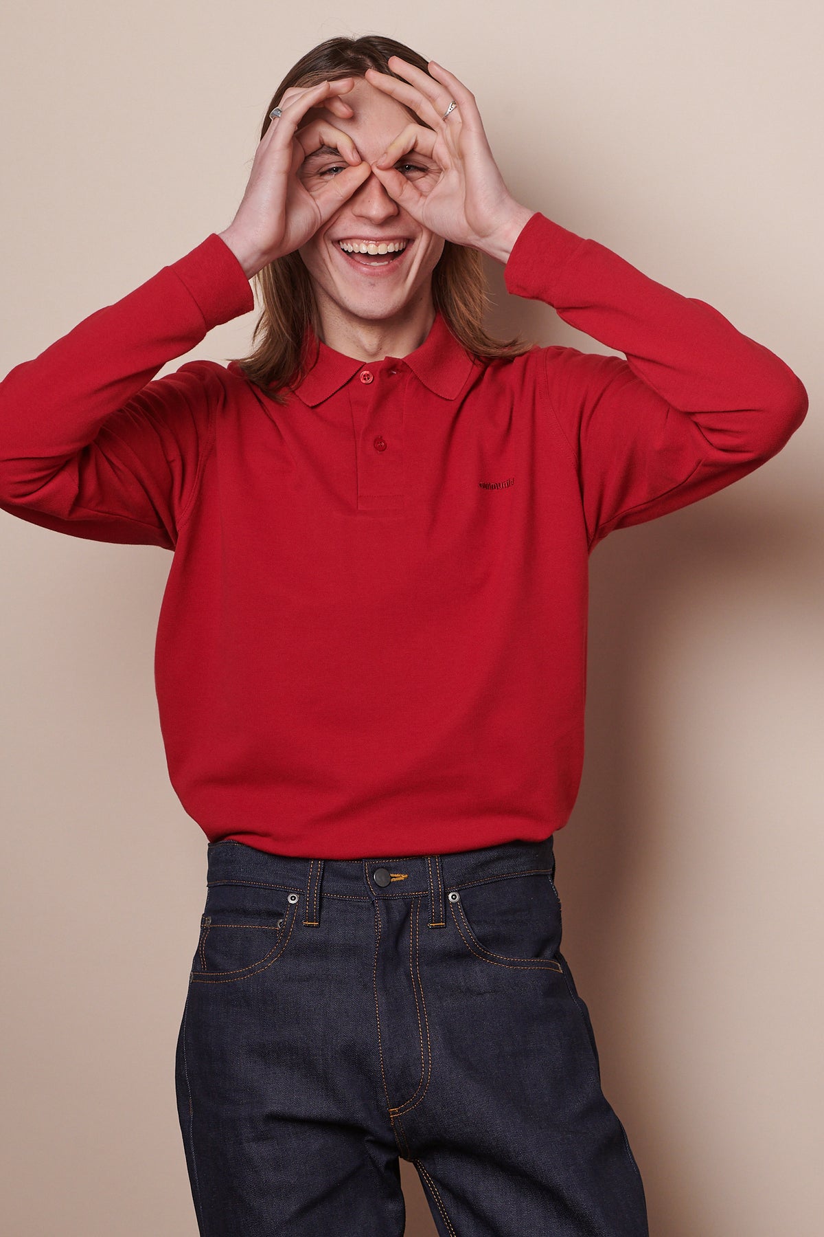 Thigh up image of white male wearing long sleeve polo shirt in crimson with arms up, holding hands in OK gesture up to both eyes. Shirt tucked into indigo jeans
