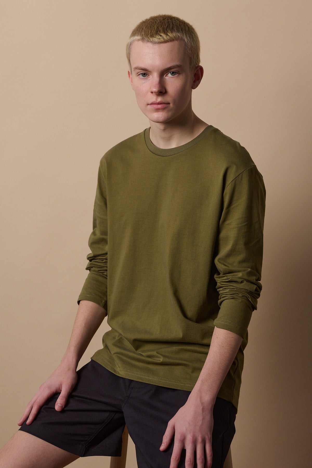 Thigh up image of white male sat on stool wearing long sleeve t shirt in olive with sleeves pushed up the arms.