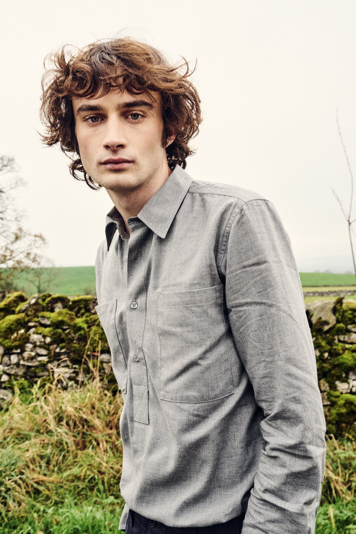 Man wearing a light gray shirt standing outdoors with greenery in the background