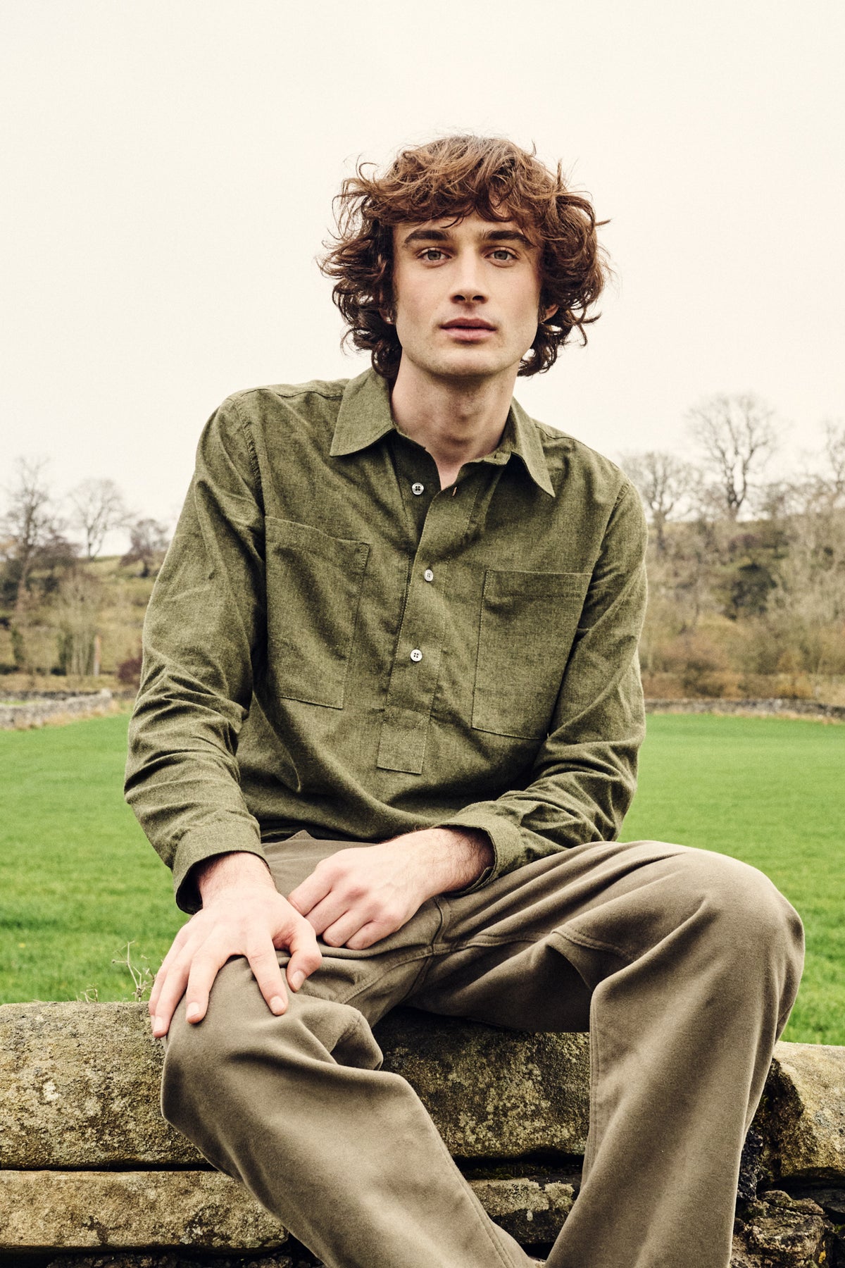 Man sitting on a stone wall in a field wearing a green shirt and olive green moleskin jeans.